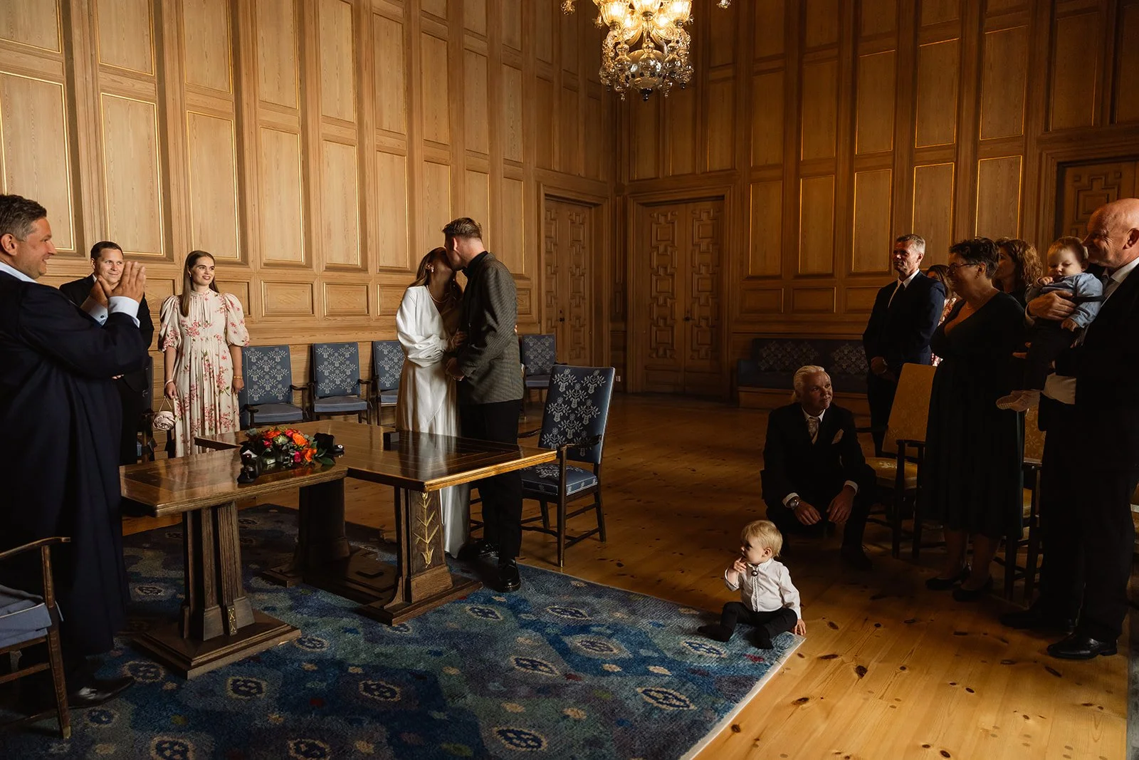 Et bryllupsbilde av en seremoni med en brud og en brudgom som kysser hverandre foran en gruppe gjester i et rom med trepanelvegger og en lysekrone. Det er en liten gutt som sitter på gulvet ved siden av et teppe, og flere voksne som ser på seremonien