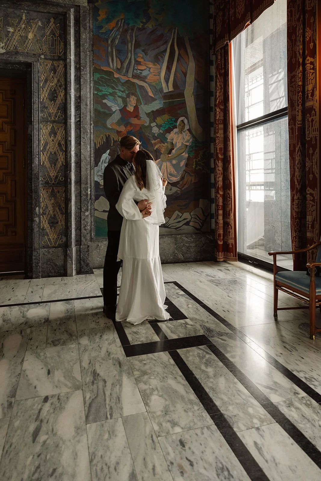 Et par som kysser hverandre i et rom med store vinduer, hvite gardiner og fargerike veggmalerier.