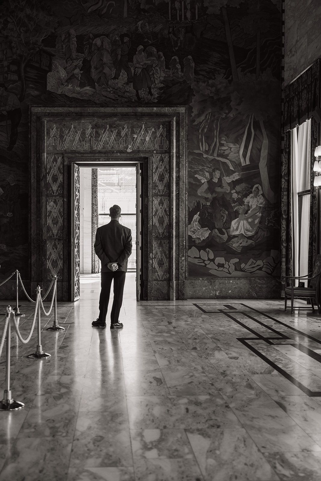 En mann i dress står med ryggen til, og ser ut av et vindu i et rom med marmorgulv og store veggmalerier.