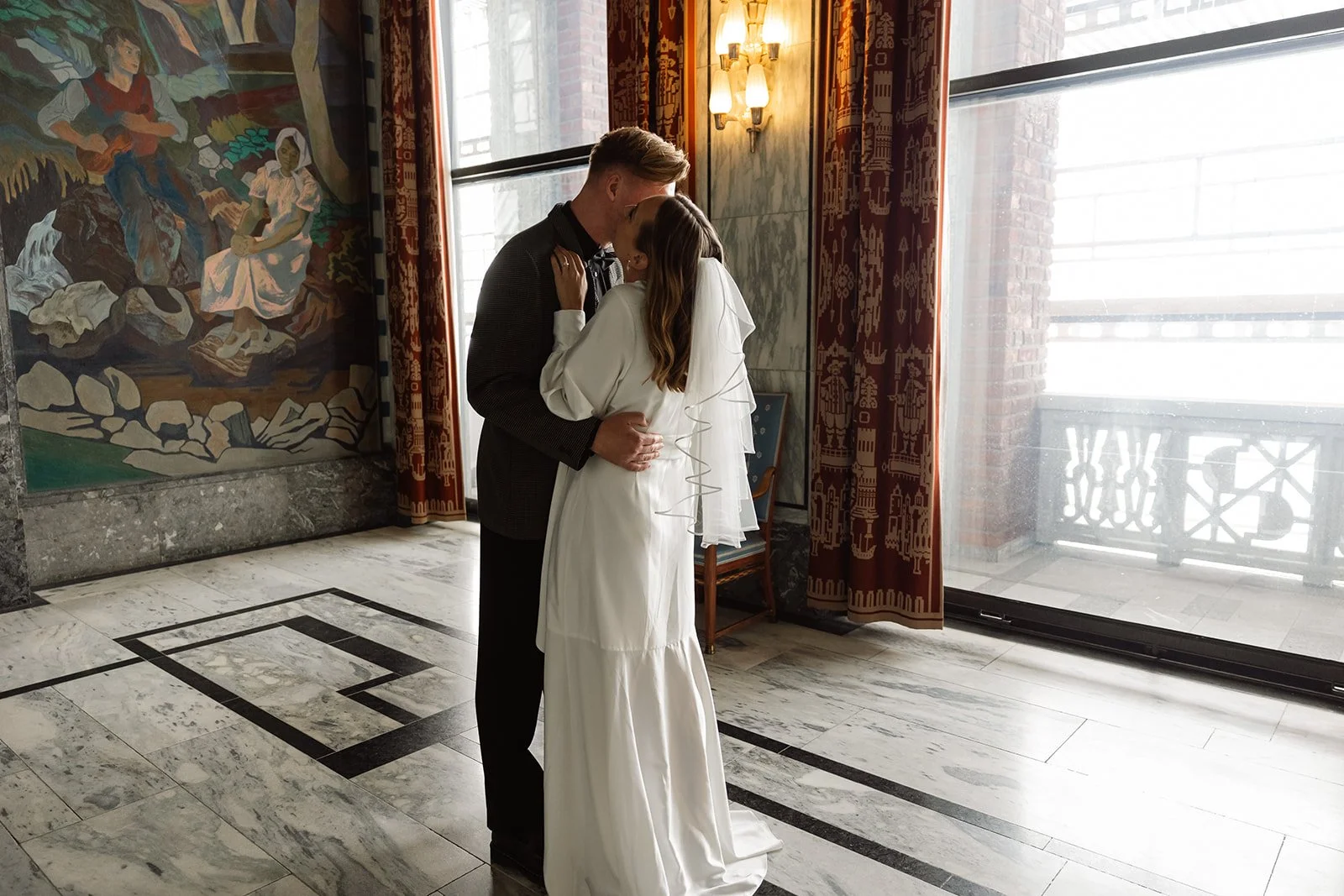 Et brudepar kysser i et rom med store vinduer, kunst på veggen, og teppe med tradisjonelle mønstre.