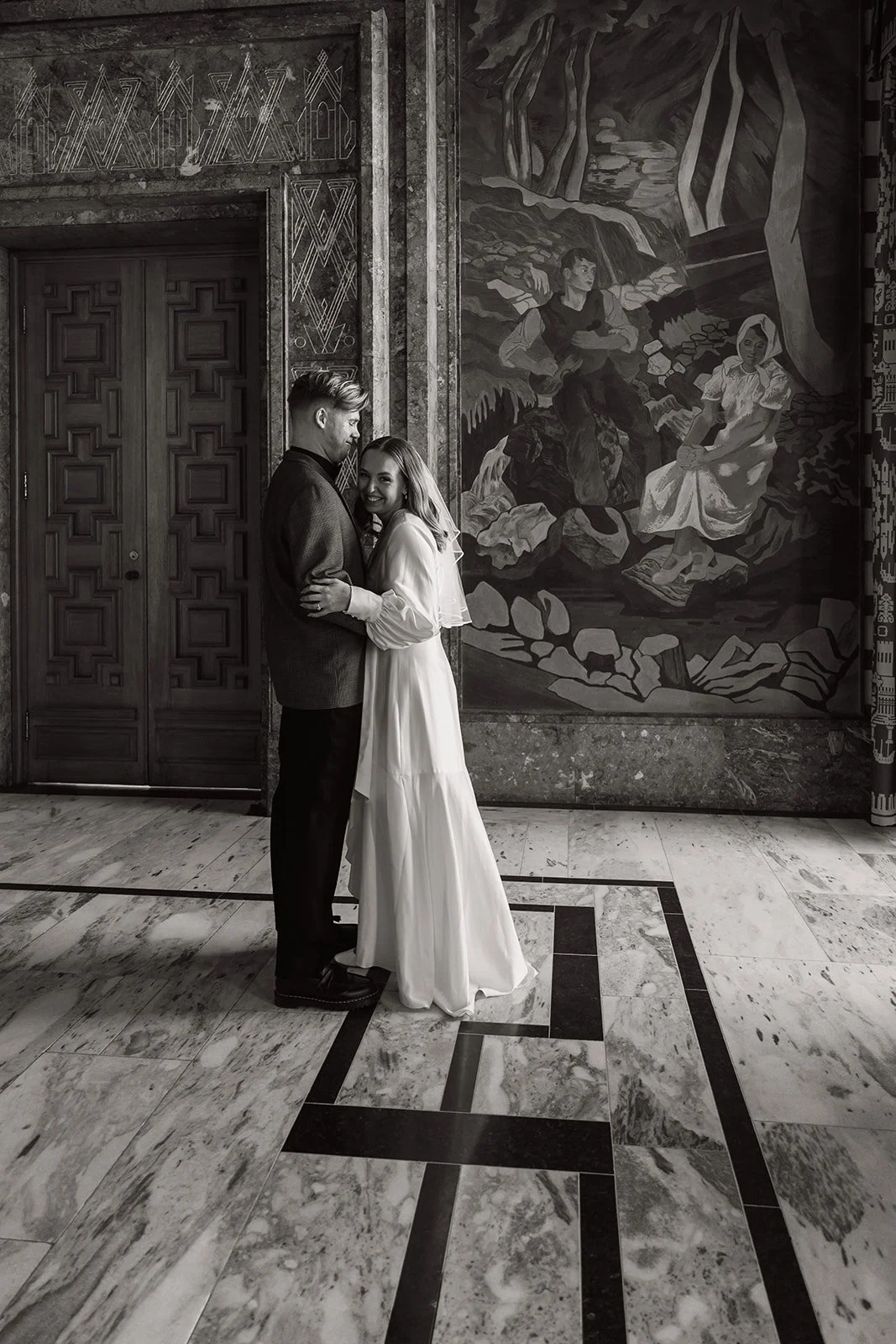 Et lykkepar av et par står sammen i en elegant innendørs setting, omgitt av kunst og arkitektoniske detaljer.