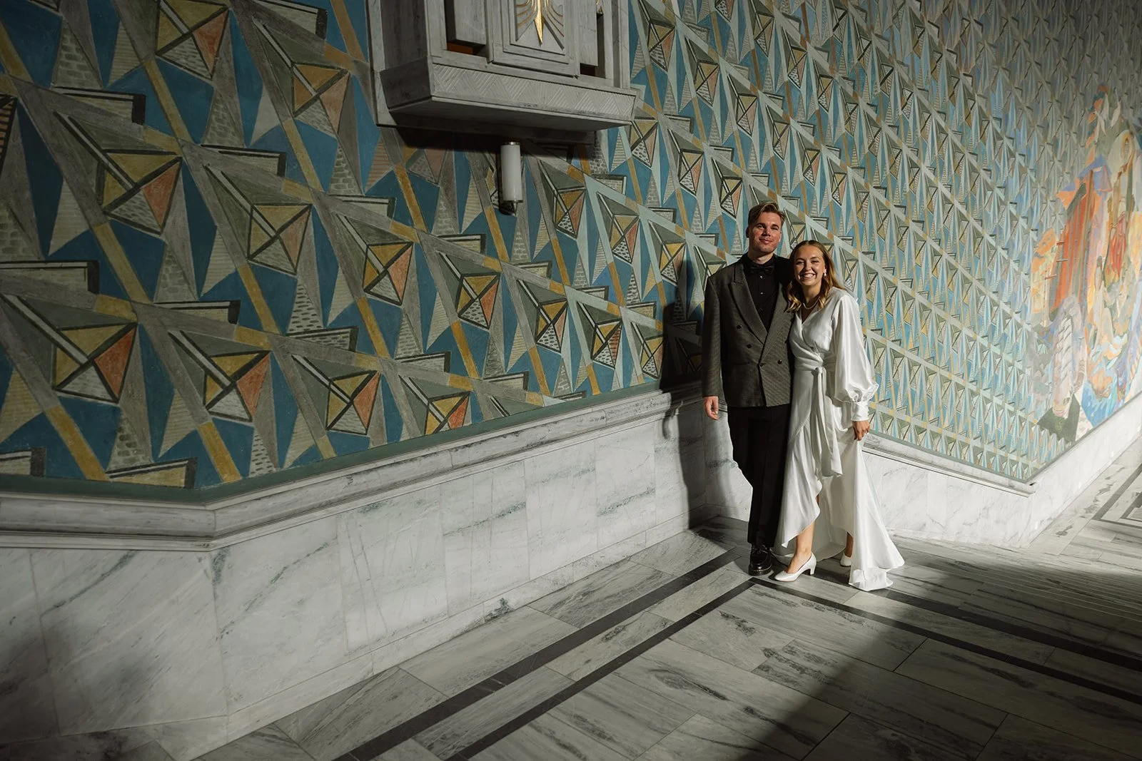 To personer står kledd i formelle antrekk og smiler foran en fargerik vegg med geometrisk kunstverk. De står på en stein- eller marmorgulv.