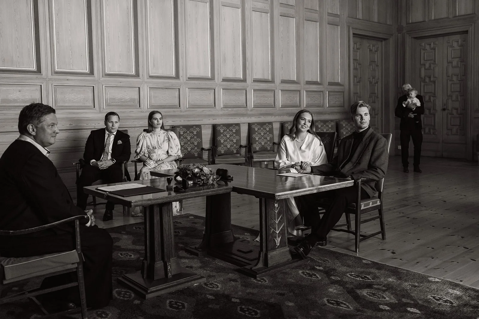 Gruppe mennesker i et formelt møte i et rom med trepaneler på veggene, noen sitter rundt et trebord, to personer står i bakgrunnen med en baby.