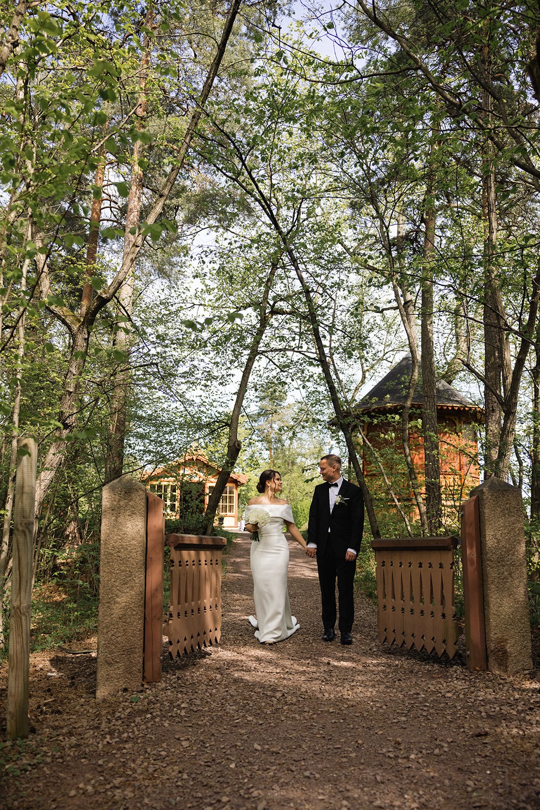 Et brudepar som går hånd i hånd gjennom en åpen port inn i en skog. Bruden bærer en hvit kjole og holder en bukett, mens brudgommen er i sort dress med sløyfe. I bakgrunnen er det små trehus omgitt av grønne trær.