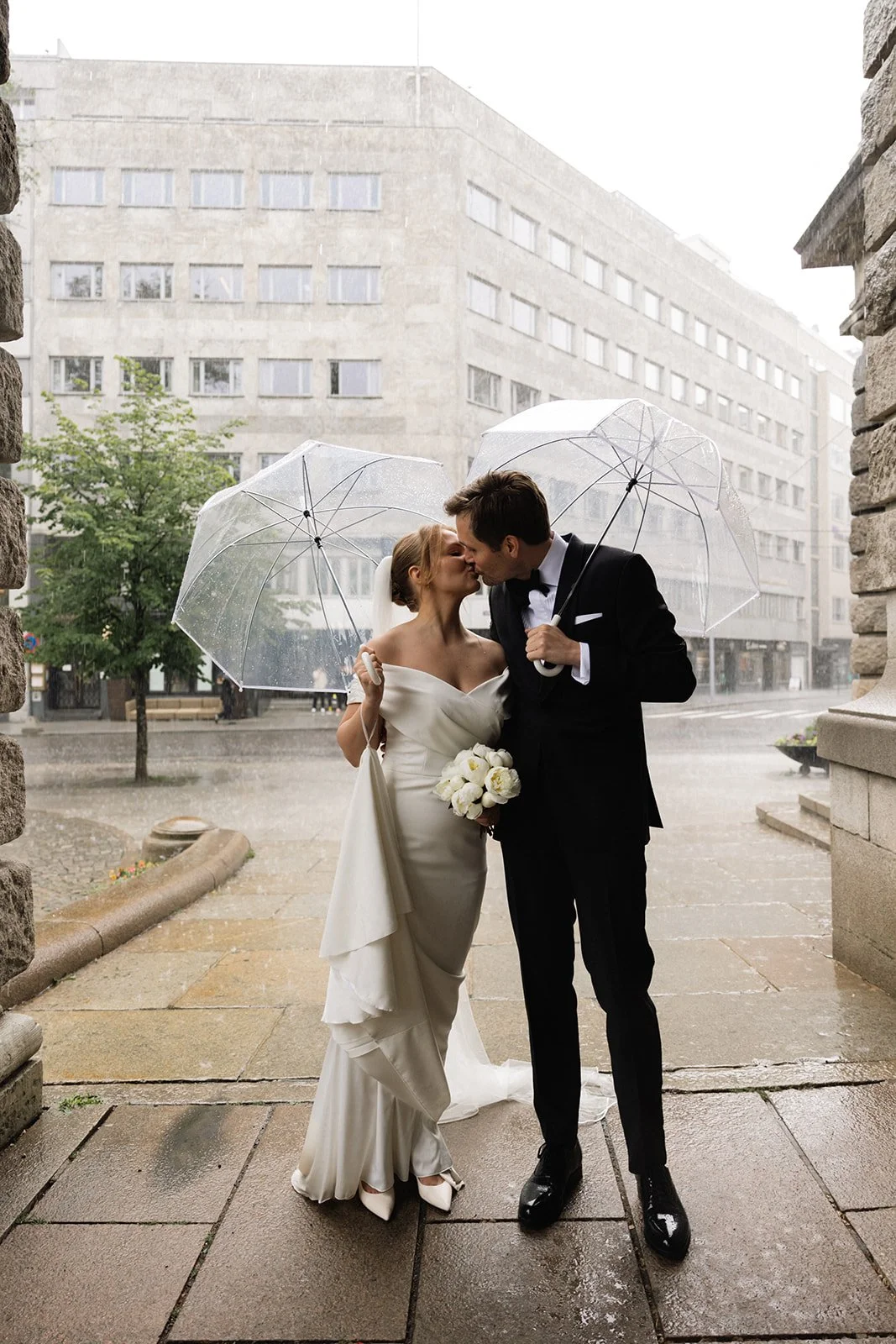 Et ektepar på bryllupsdag kysser under en åpenstående paraply i regnet foran en bygning.