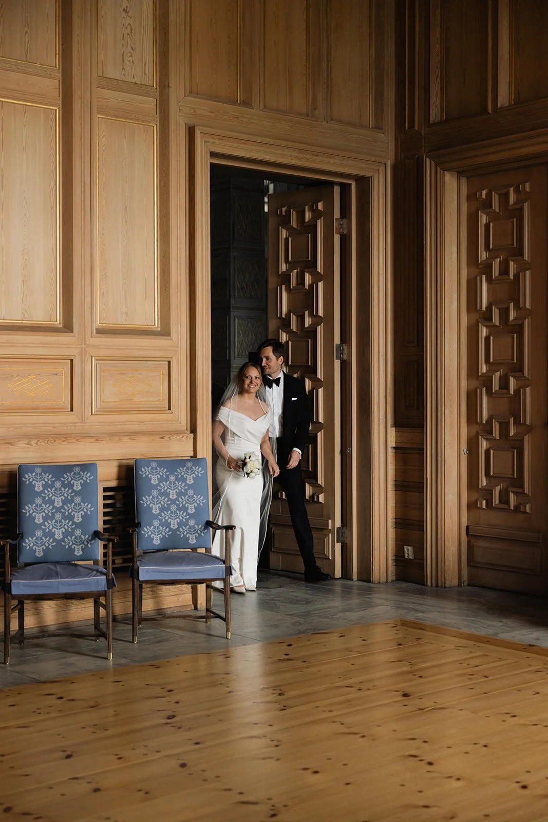 En brud og brudgom står i en stor, treskandinavisk rom med innfelt panel på veggene. Bruden bærer hvit kjole og holder en liten blomsterbukett, mens brudgommen har på seg en sort dress og hvit skjorte.