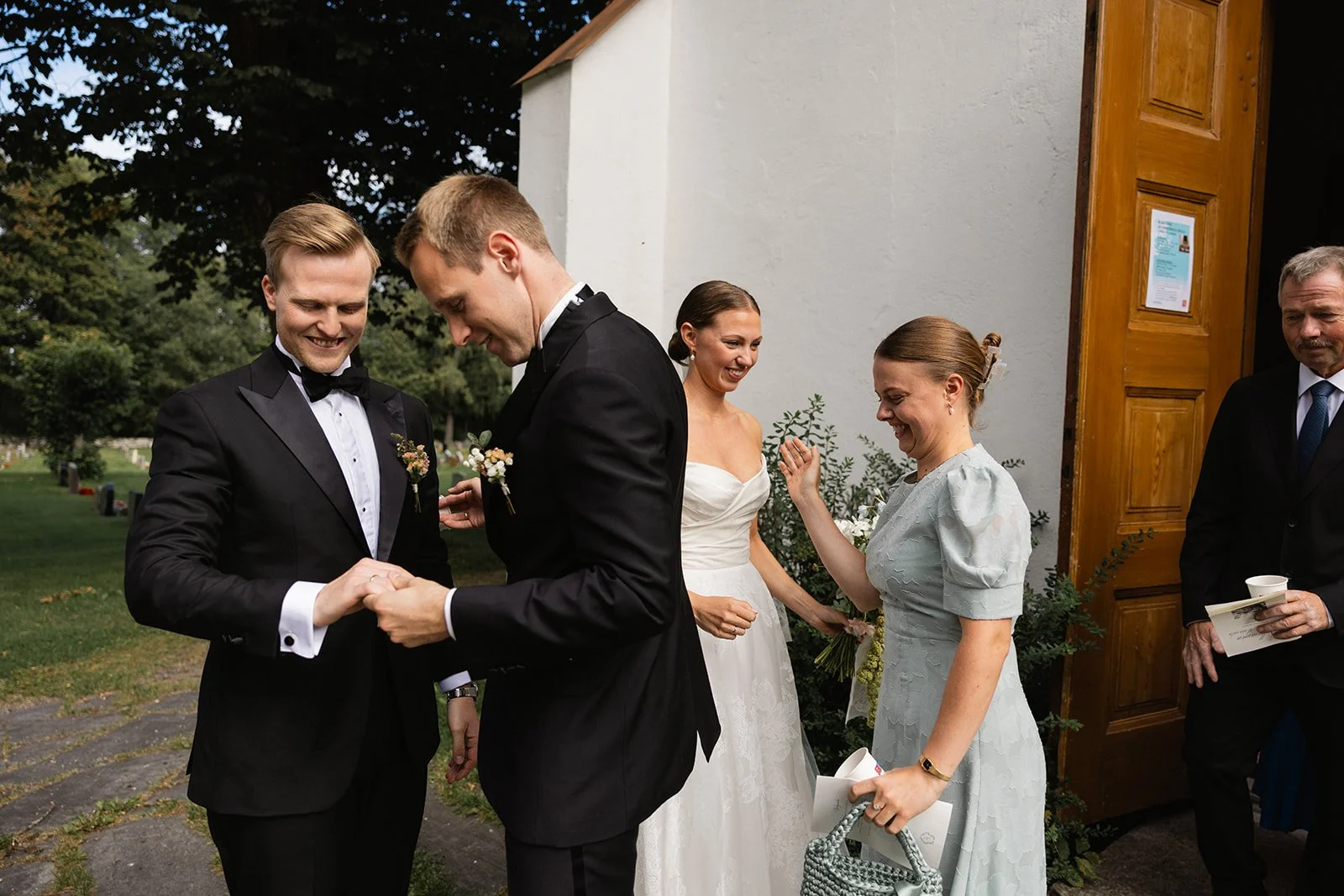 To menn i formelle klær holder hender foran en kirke, med tre kvinner i kjoler som smiler og snakker.