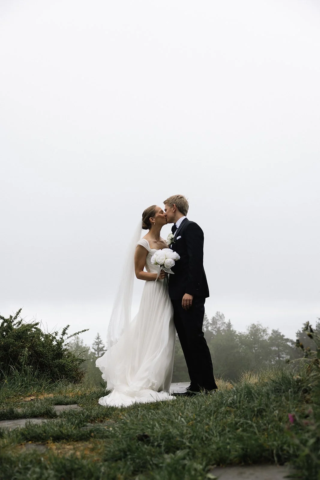 Et brudepar kysses utendørs på en grussti omgitt av grønt vegetasjon under en overskyet himmel.