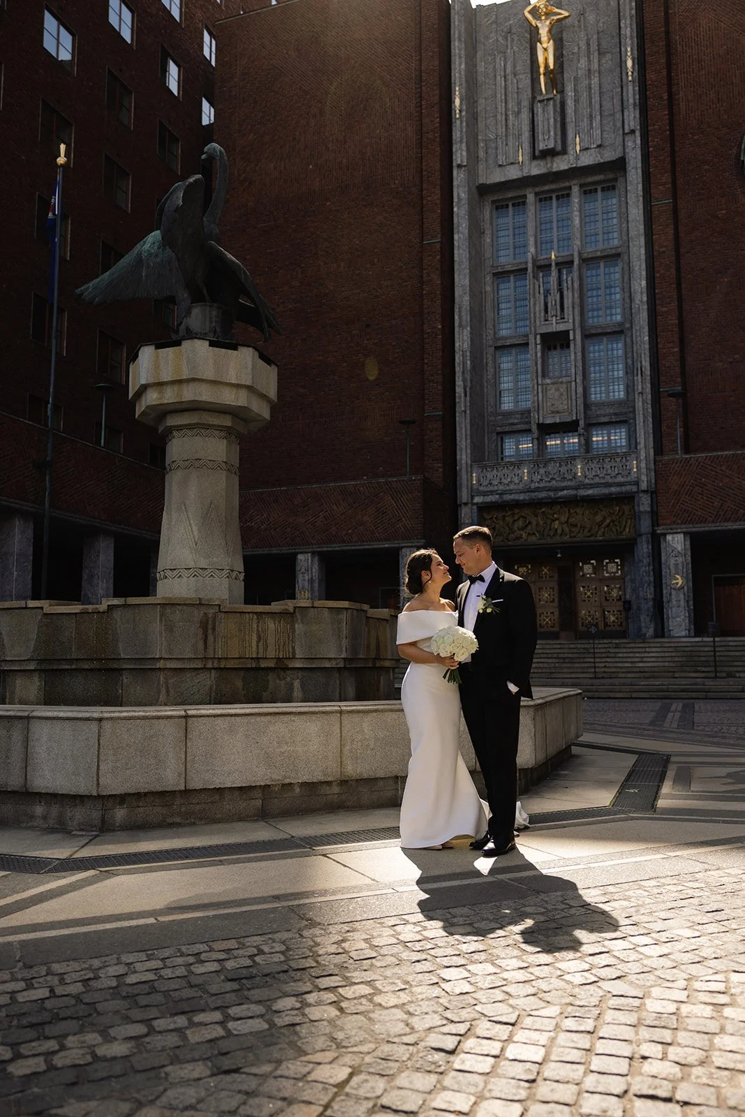 Et nygift par står foran en skulptur utenfor en bygningsmur. Damen har på seg en hvit kjole og holder en blomsterbukett, mens herren er i svart dress og slips.