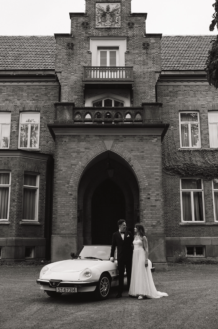 Et brudepar står foran en hvit Alfa Romeo Spider utenfor en stor mursteinsbygning med trapper og balkonger.