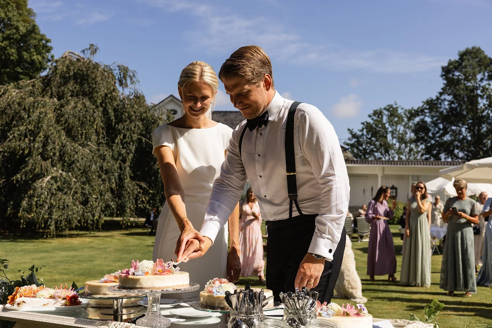 En brud og brudgom klemmer en brytekake under en utendørs bryllupsseremoni med gjester i bakgrunnen.