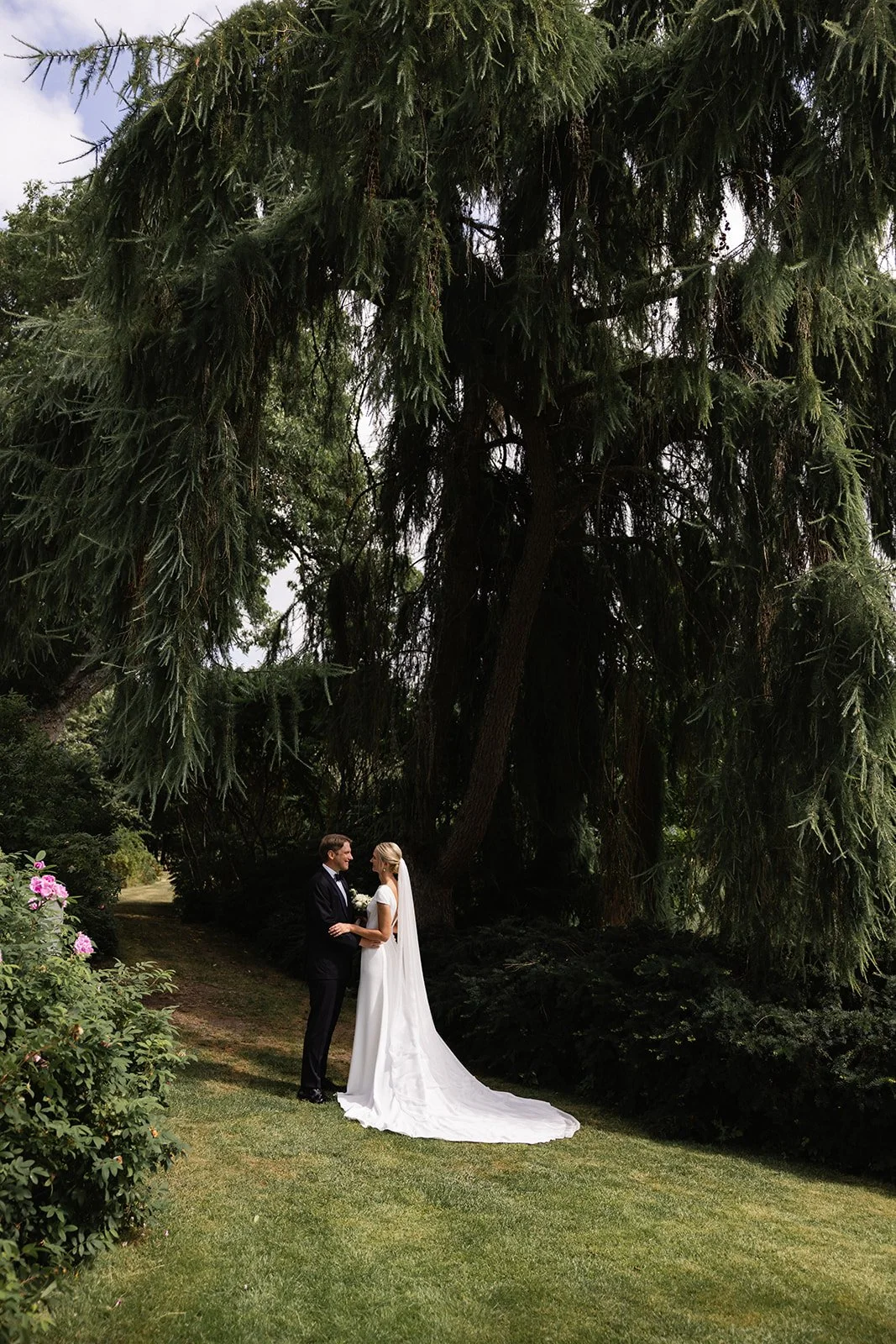Et par i brudekjole og dress holder hender foran et stort tre i en hage, omgitt av grønn vegetasjon og blomster.