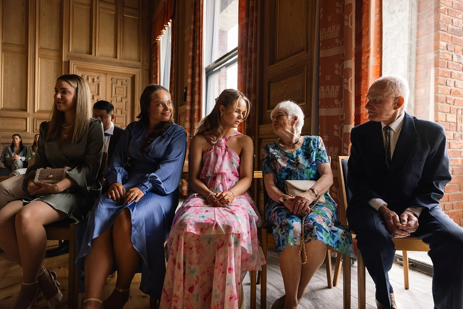 En gruppe mennesker sitter i en rekke i et rom med store vinduer og trevegger, iført fine klær, antagelig til en seremoni eller seremoniell begivenhet.