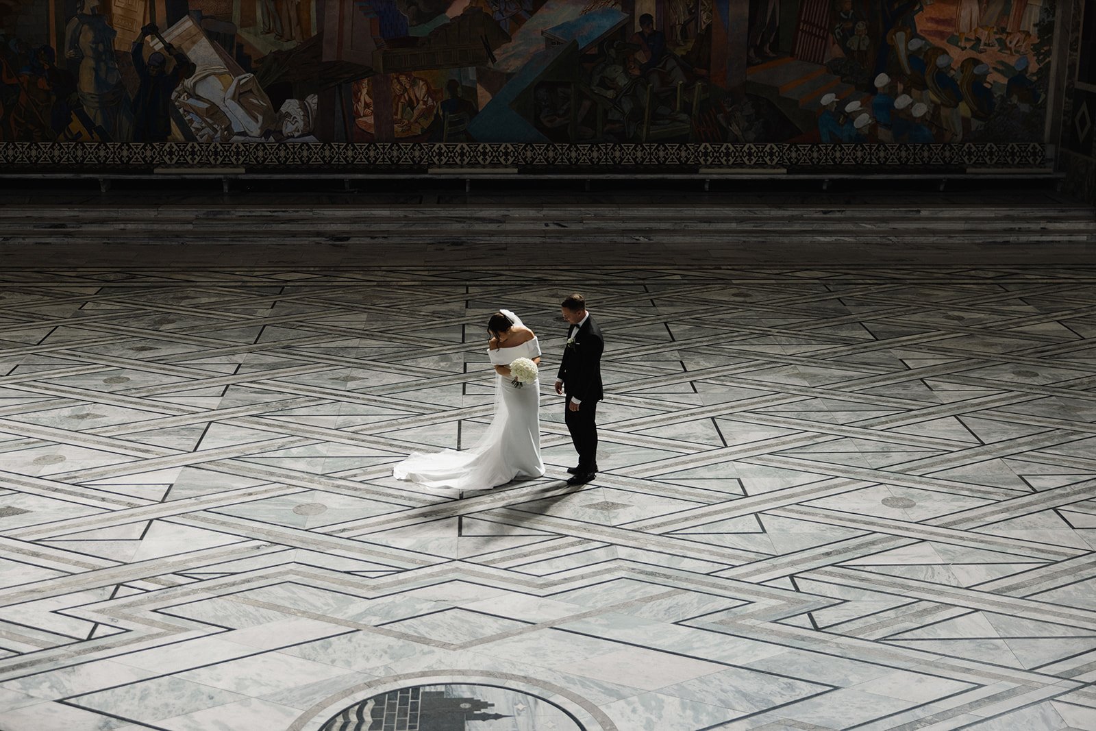 En brude og brudgom står midt på et stort, tomt, mønsterdekket gulv innendørs, med en stor dekorativ vegg i bakgrunnen. Bruden bærer en hvit kjole og holder en blomsterbukett, mens brudgommen er i dress.