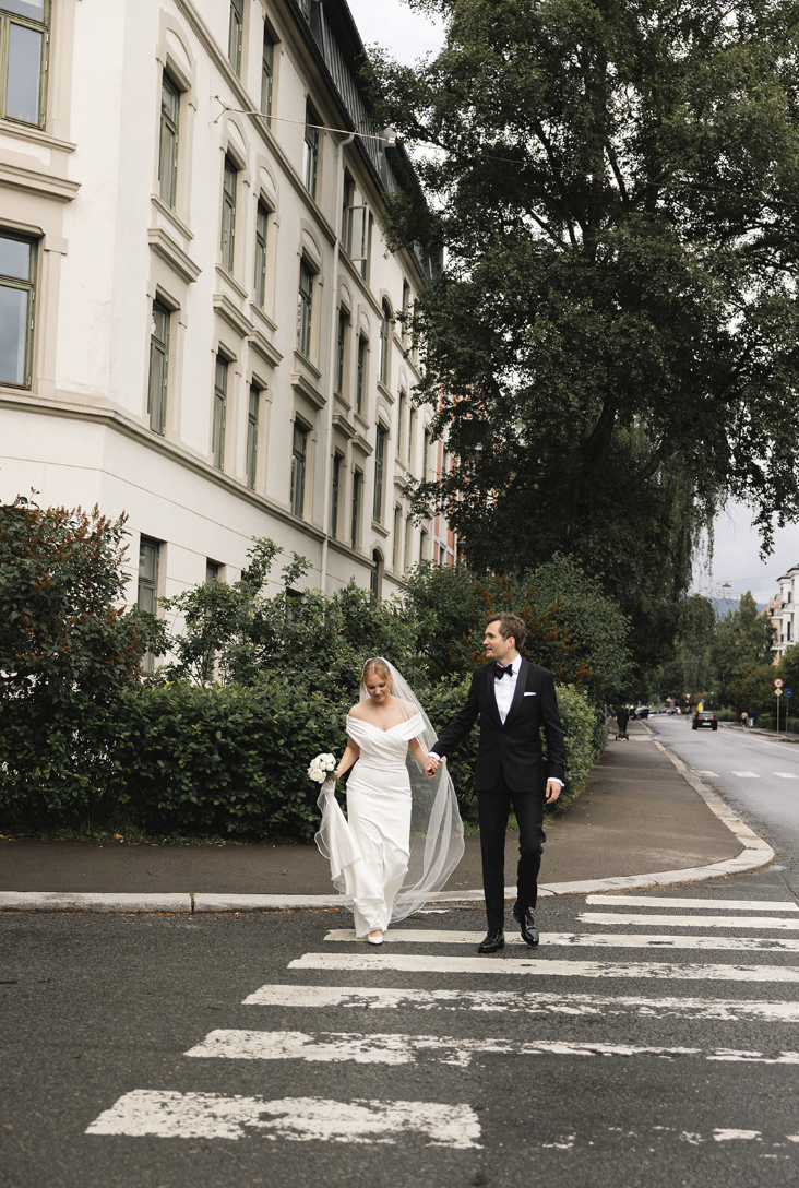 En brud og en forlover går hånd i hånd over et fotgjengerovergang. Bruden har på seg en hvit brudekjole og holder en bukett med hvite blomster. Mannen har på seg en svart smokingsjorte og slipse. De går langs en gate med trær og bygårder.