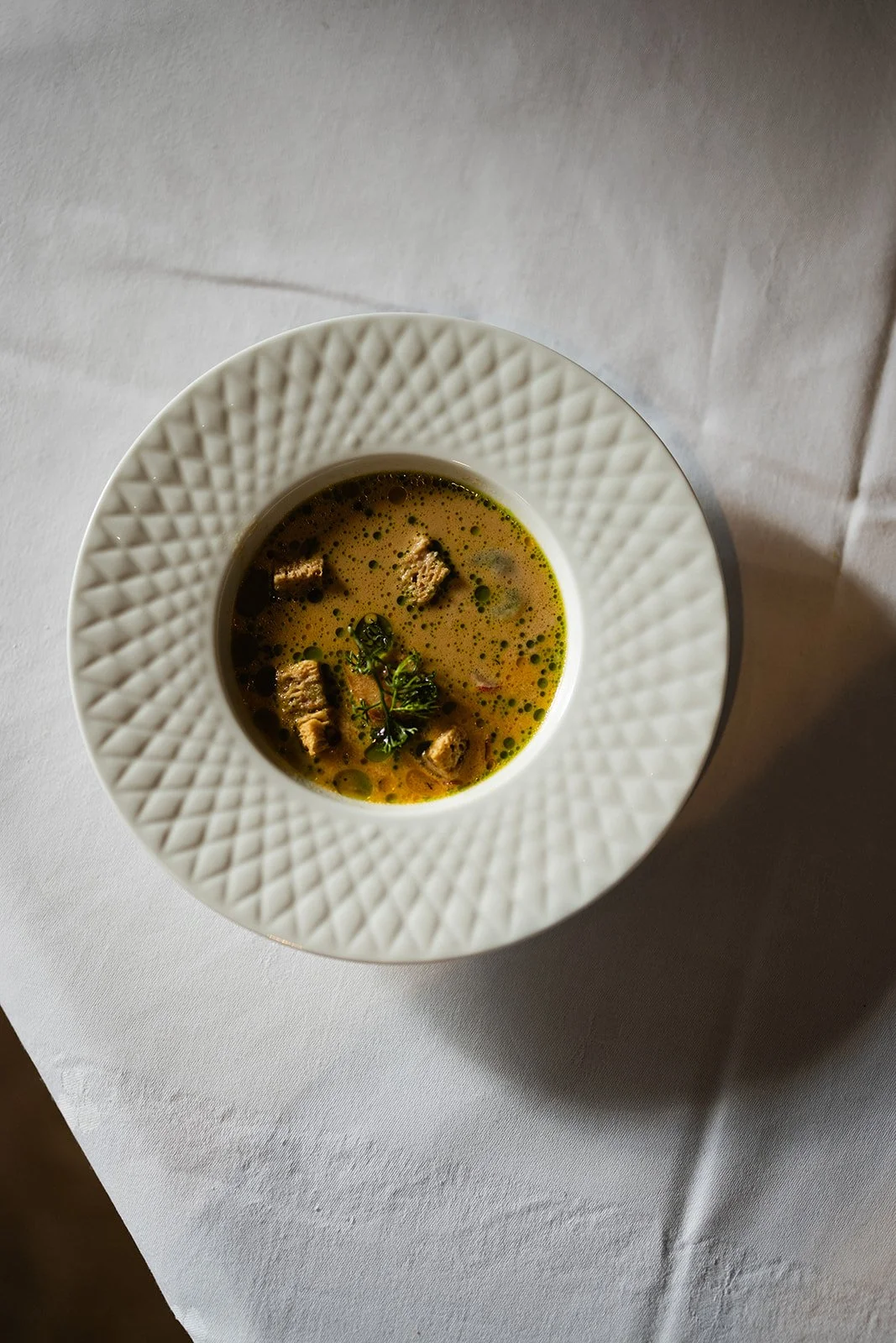 En tallerken med suppe med små kjøttbiter og urter, servert på en hvit tallerken med et mønster rundt kanten, plassert på en hvit duk.