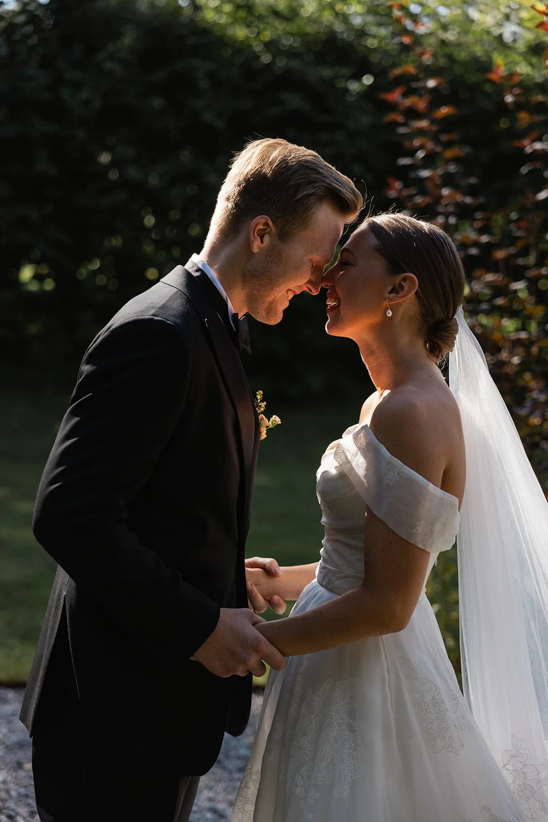 Et brudepar står ansikt til ansikt, holder hender, i naturen med sollys som skinner mellom trærne, smiler og nyter øyeblikket.