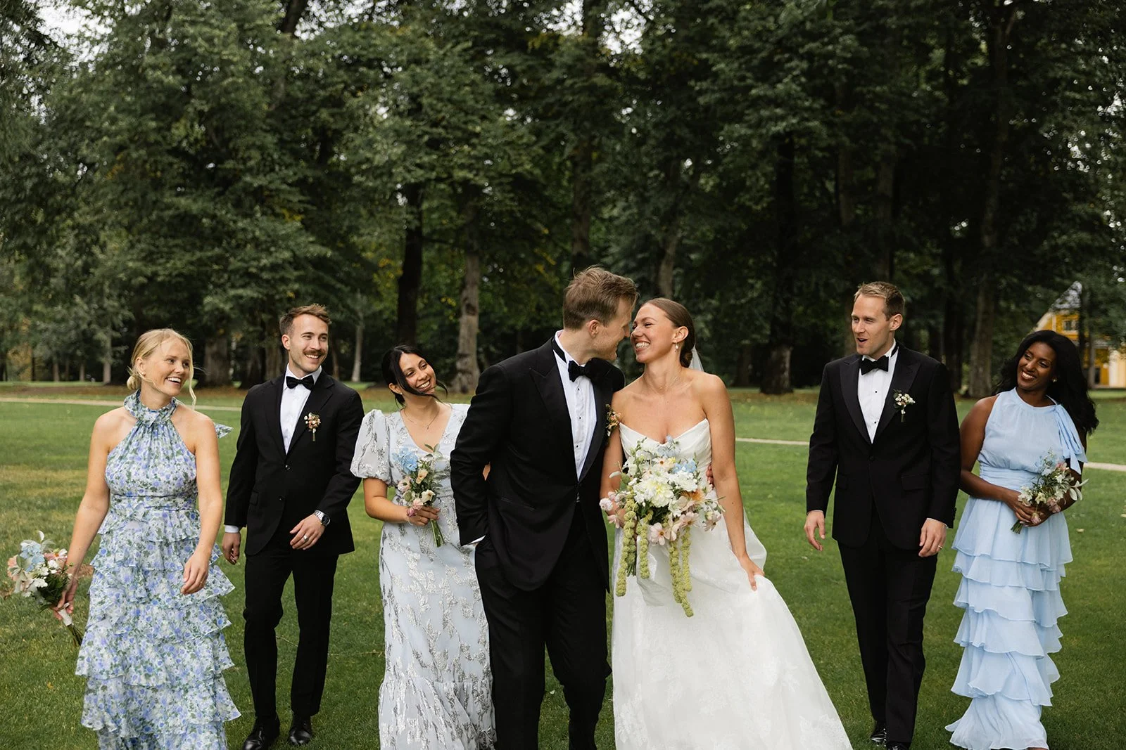 Brudepar smiler og går hånd i hånd, omgitt av brudepiker og forlovere i en grønn park.