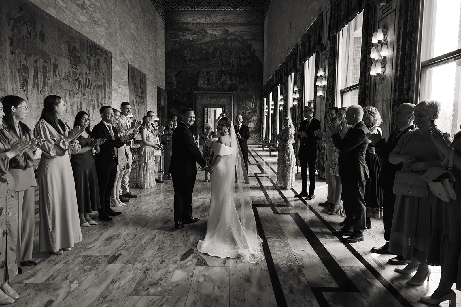 Et svart-hvitt bilde av et bryllup med brudeparet i midten, hånd i hånd, omgitt av gjester som klapper og smiler. Bilde tatt innendørs i en elegant sal med store vinduer, gardiner og veggmalerier.