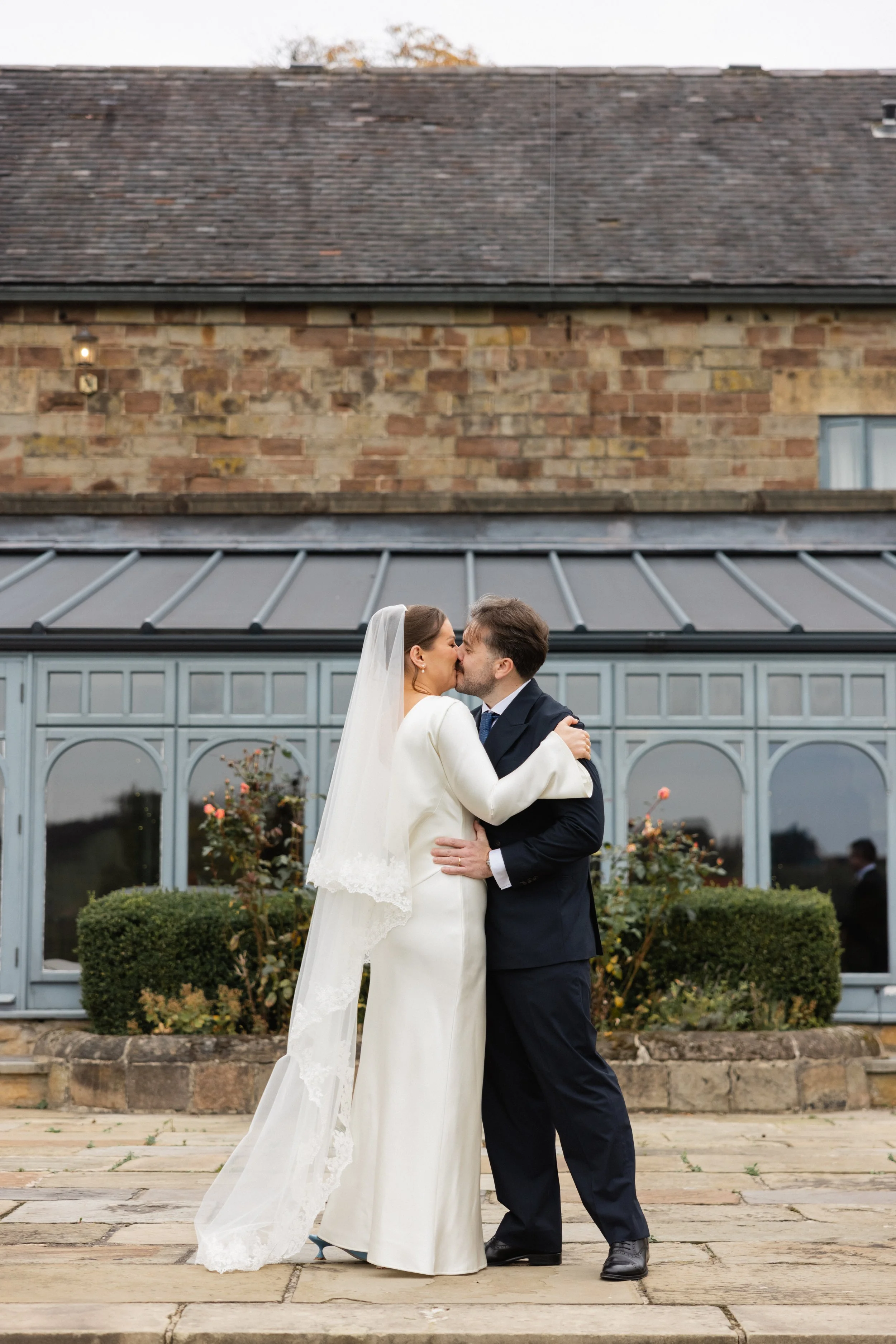 Et nygift par kysser utenfor en gammel murbygning, kledd i bryllupsantrekk, med planter og blomster i bakgrunnen.
