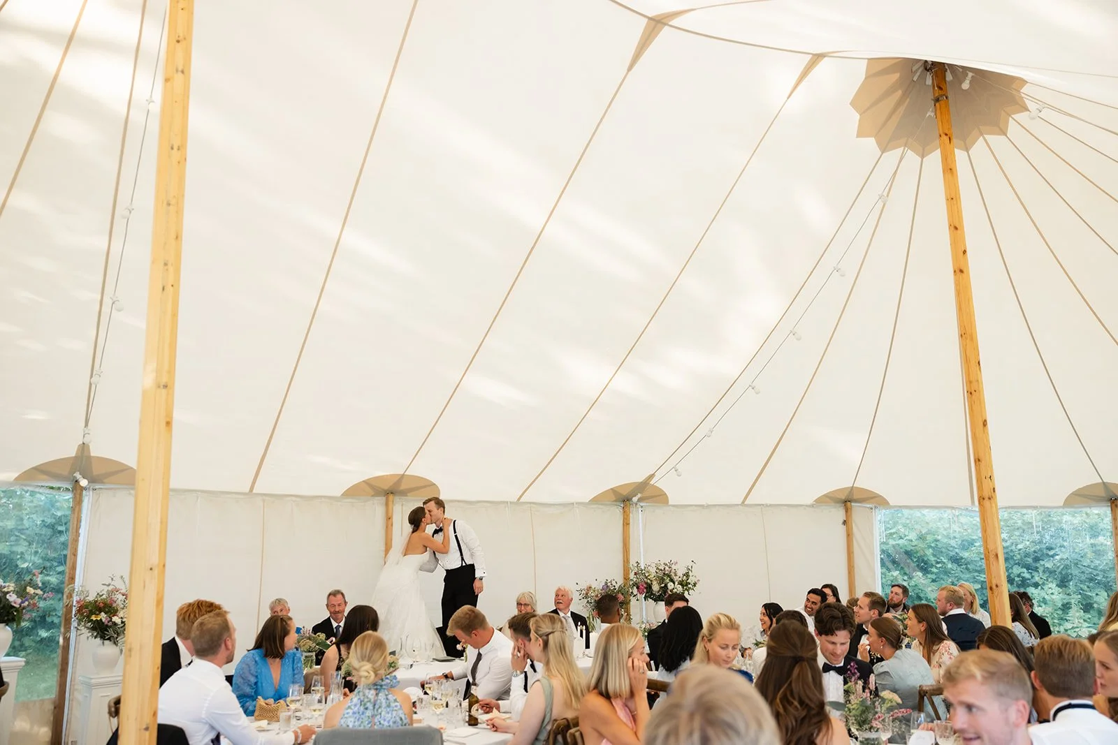 Brudepar kysser under en stor hvit festtelt med mange gjester sittende ved bord.