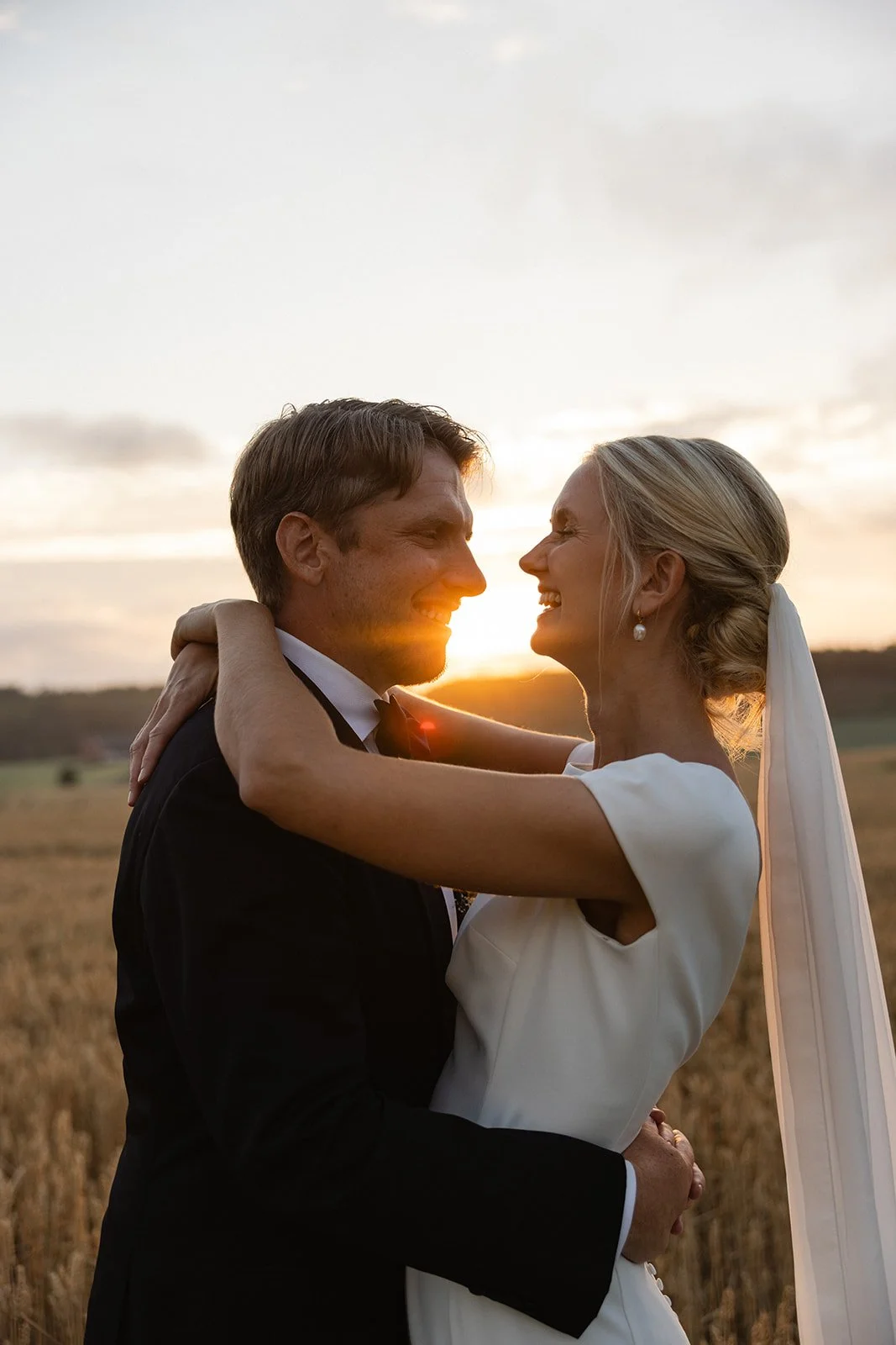 Et lykkelig brudepar klemmer hverandre i en åker under solnedgang.