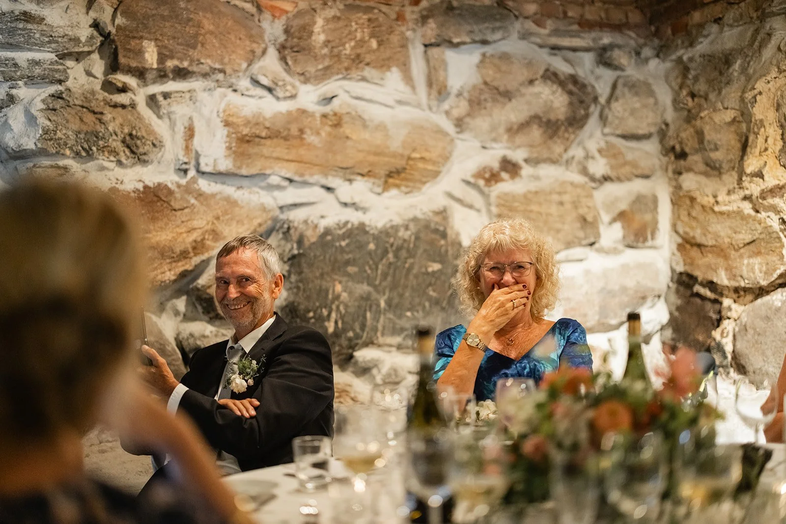 To personer som sitter ved et bord under en seremoniell anledning, med en stein- eller murvegg i bakgrunnen. Den ene er en mann i dress, den andre er en kvinne med blond, krøllete hår og briller, og de smiler og ler.