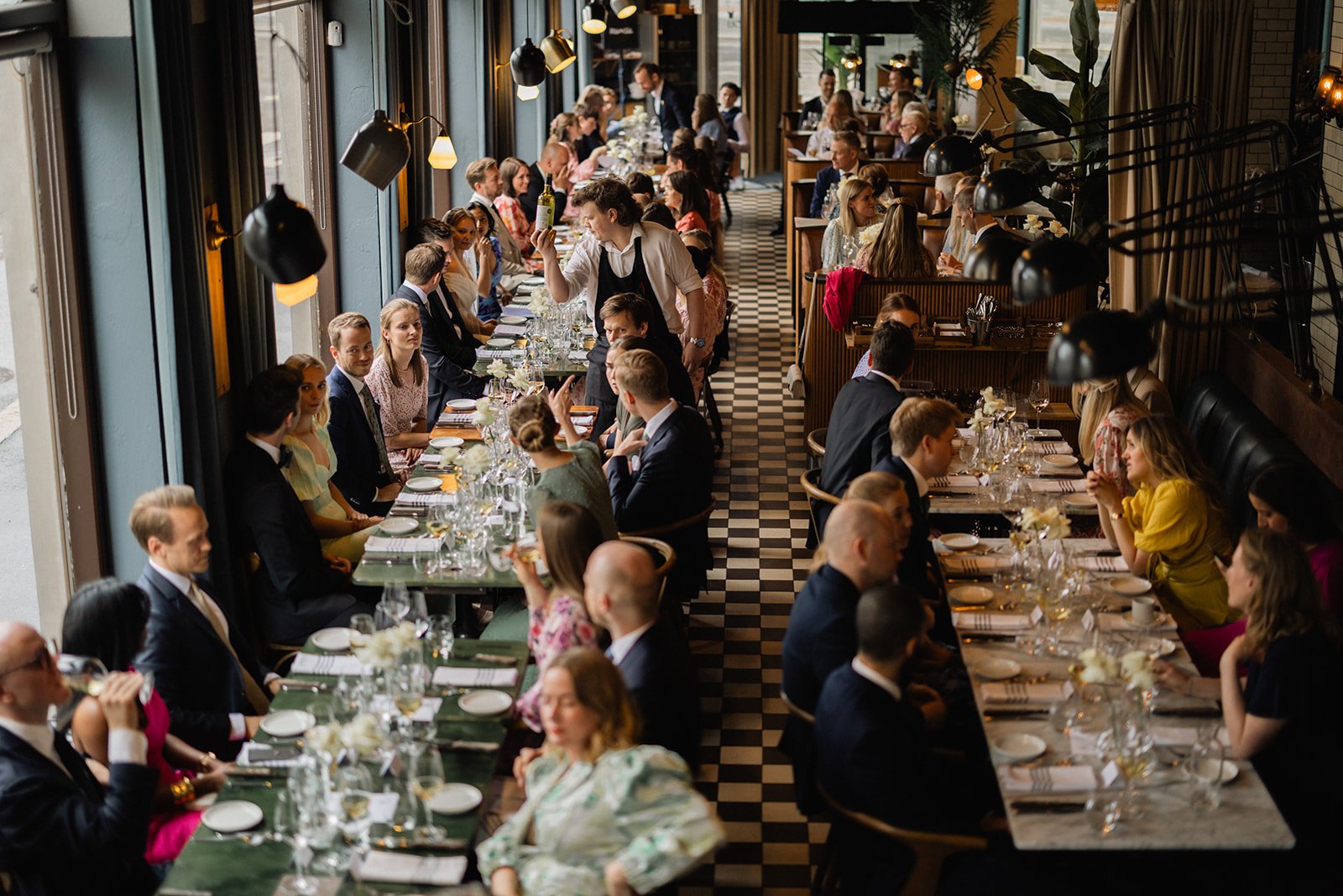 En restaurant med mange gjester som sitter ved langbord, og en servitør som bærer en flaske vin.