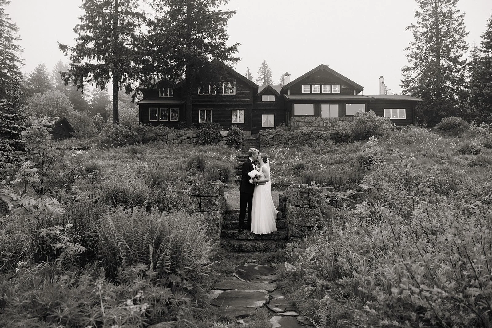 Et brudepar står kysser på en liten trapp i en hage, med en stor bolig i treverk i bakgrunnen.
