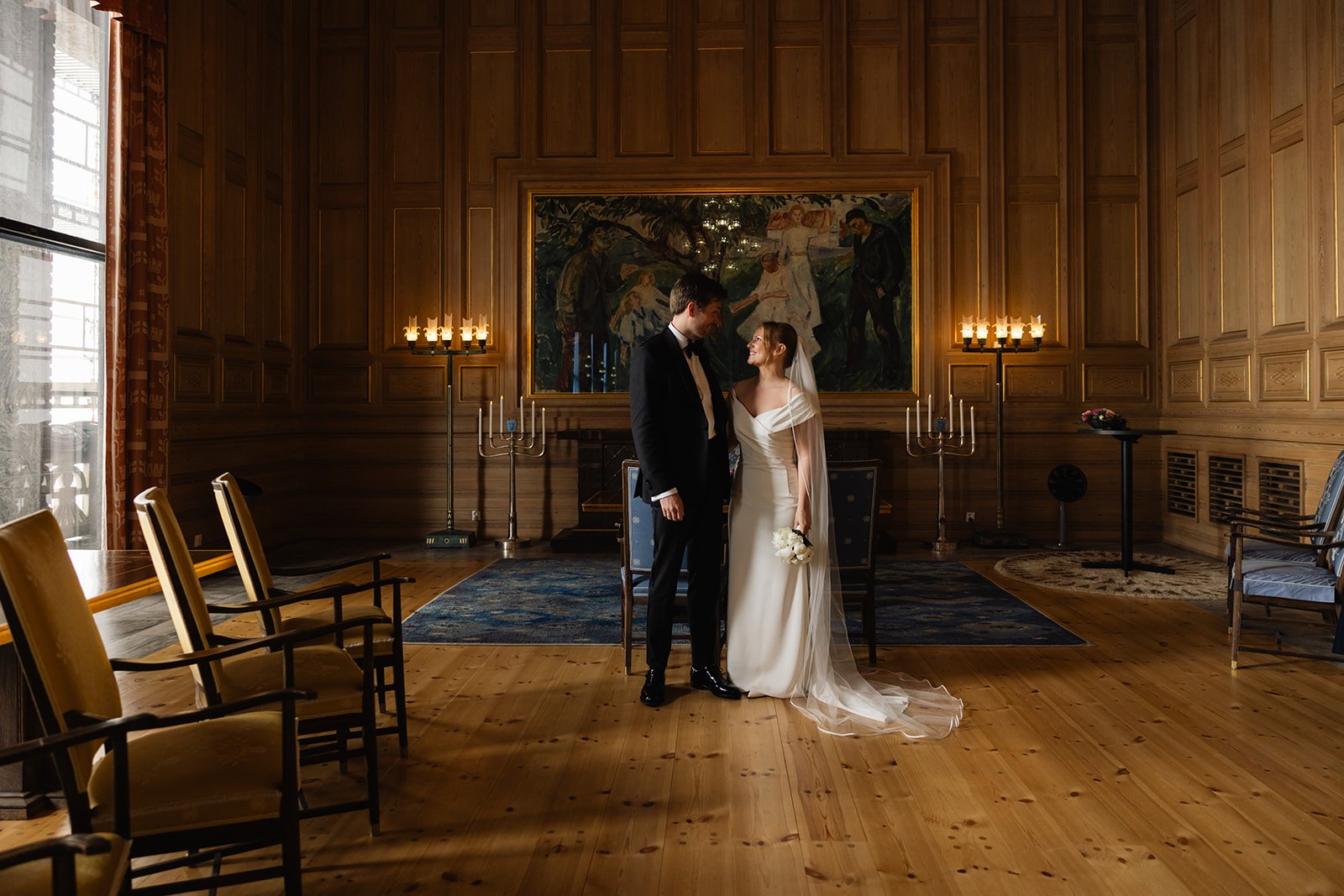 En nygifte brudepar står i et rom med tremøbler og veggpaneler, med innrammet maleri bak seg, og holder hender mens de smiler til hverandre.