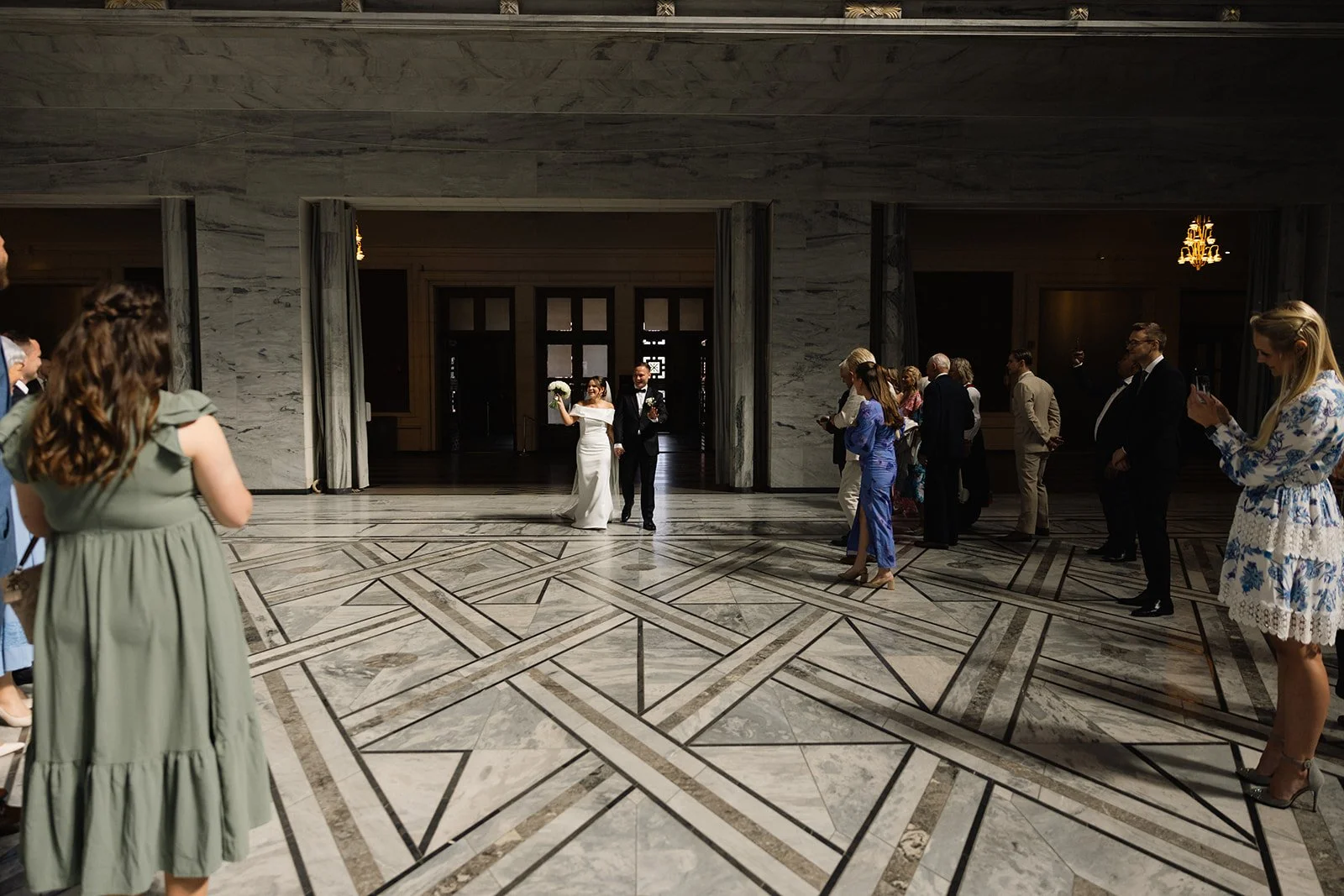 Bryllupsparet kommer inn i en stor, elegant hall, omgitt av gjester. Bruden bærer en hvit kjole og holder en blomsterbukett, mens brudgommen har på seg dress og slips.