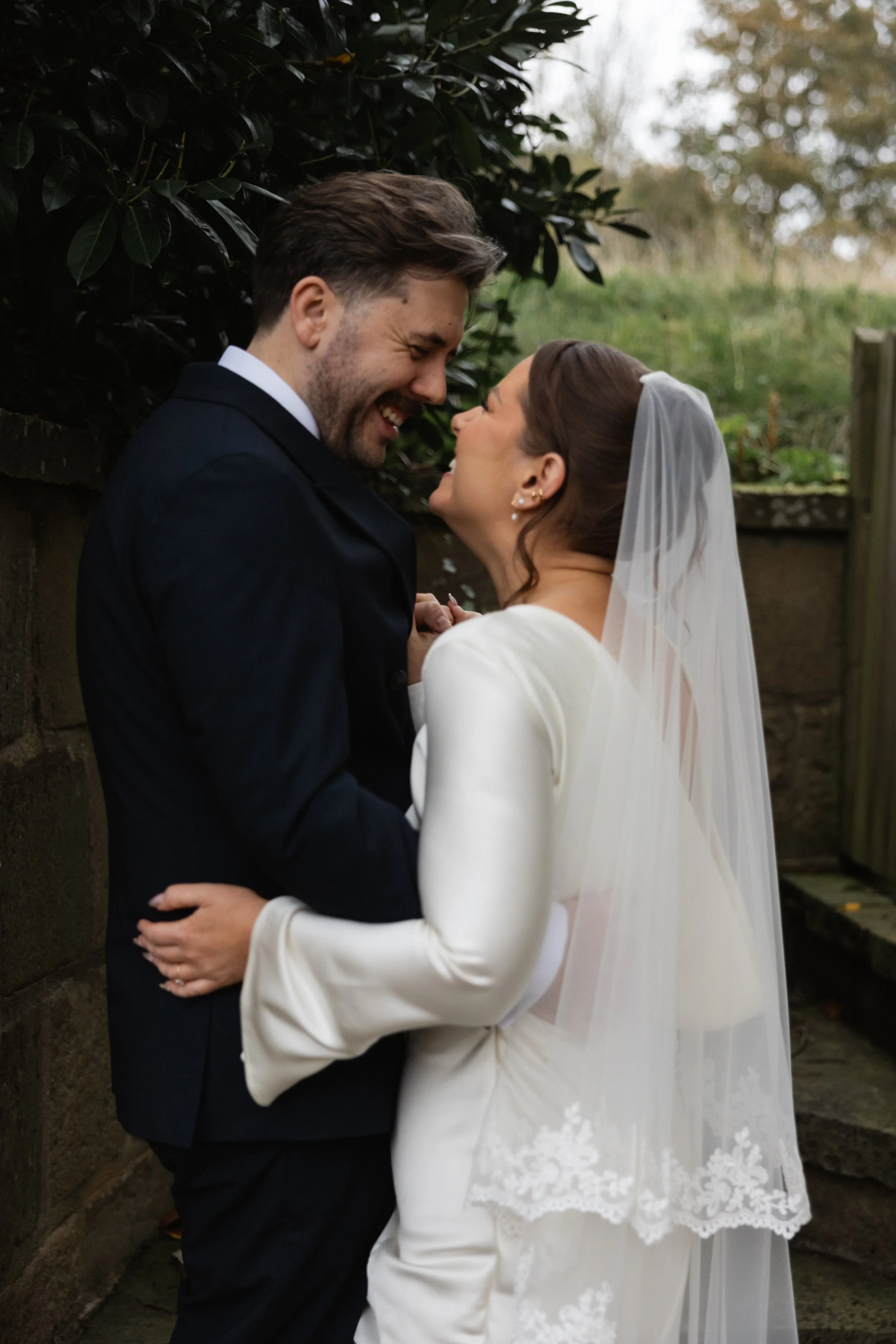 Et lykkelig nygift par som smiler og ser på hverandre, med brudekleid og dress, ute i naturen.