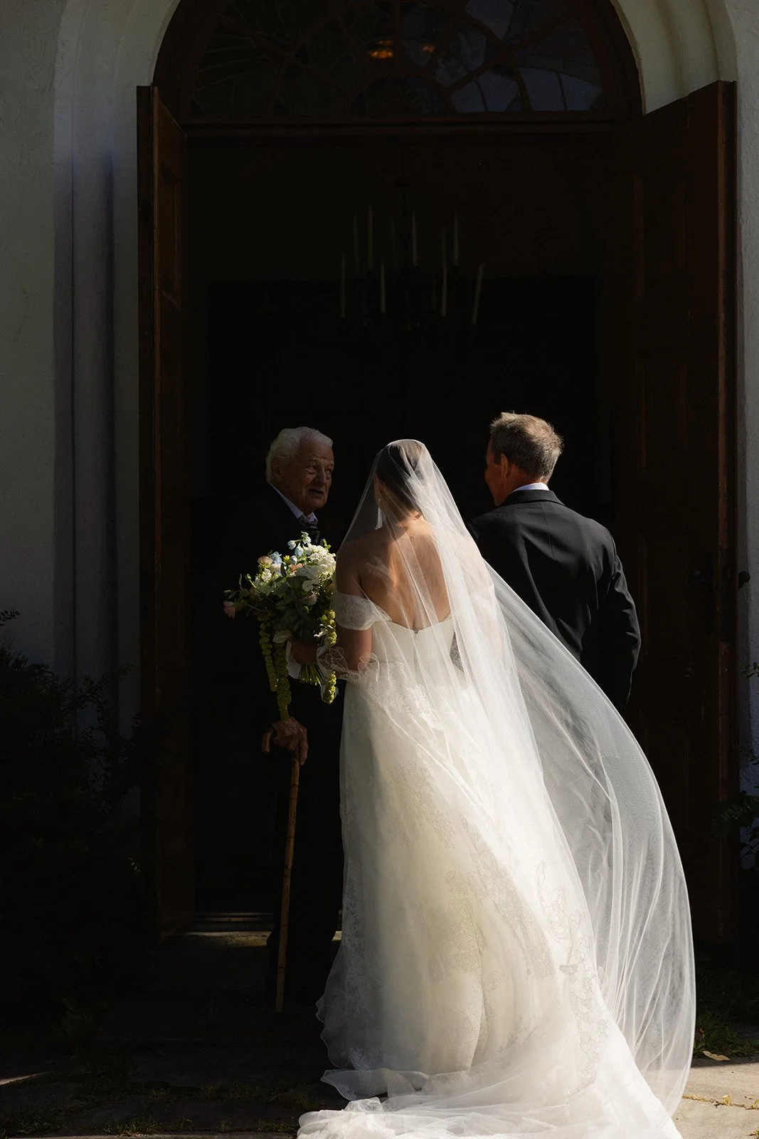 En brud i hvit kjole og slør står foran en eldre mann med blomsterbukett og en mann i dress utenfor en åpen dør, i et kirkerom