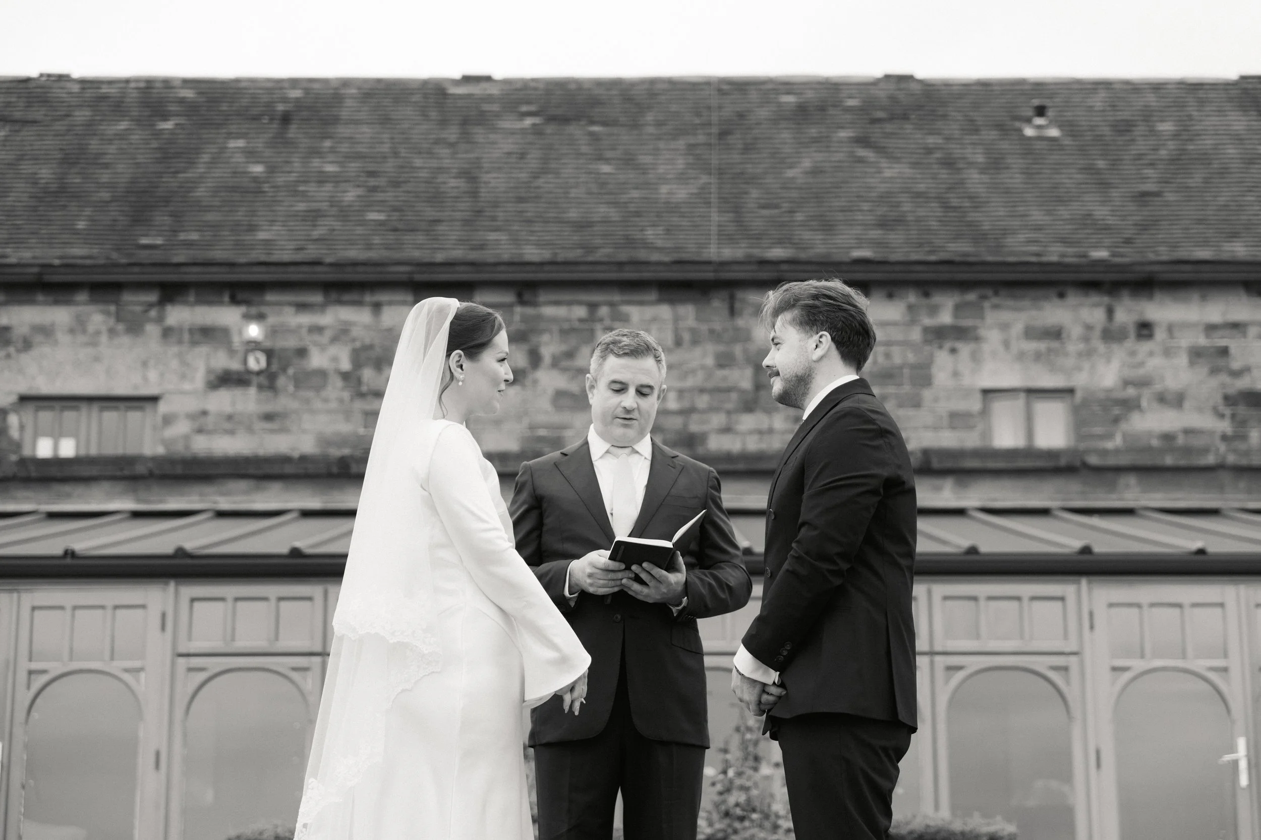 Et bryllupsseremoni med en brud og en brudgom som holder hender, med en offisiant som leser fra en bok, utendørs foran en bygning.