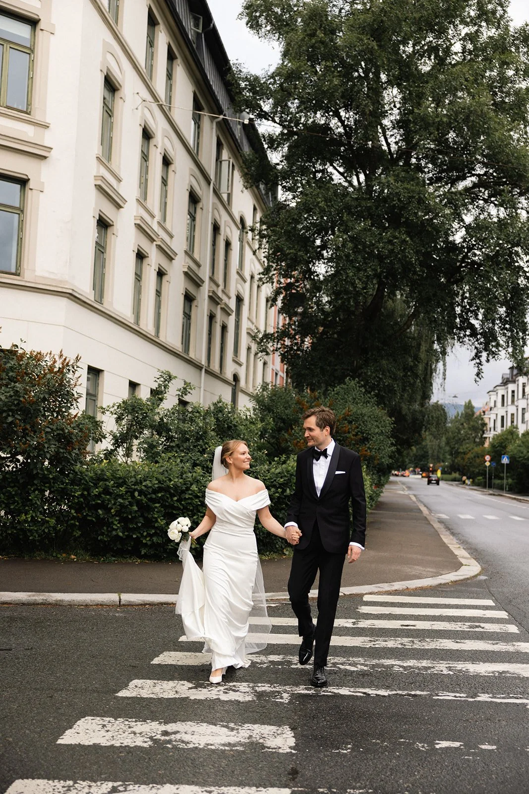 Et bryllupspar går hånd i hånd over et fotgjengerovergang på en gate, med grønne busker og bygninger i bakgrunnen.