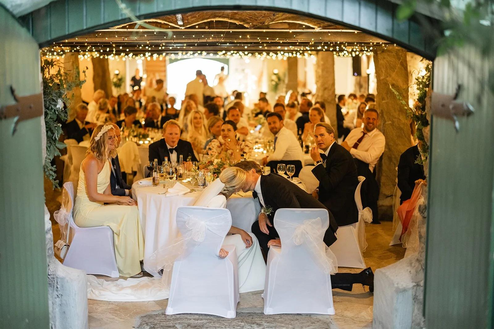 Bryllupsmiddag under tak med gjester som sitter ved bord, dekorert med lys og blomster, sett gjennom en åpen dør.