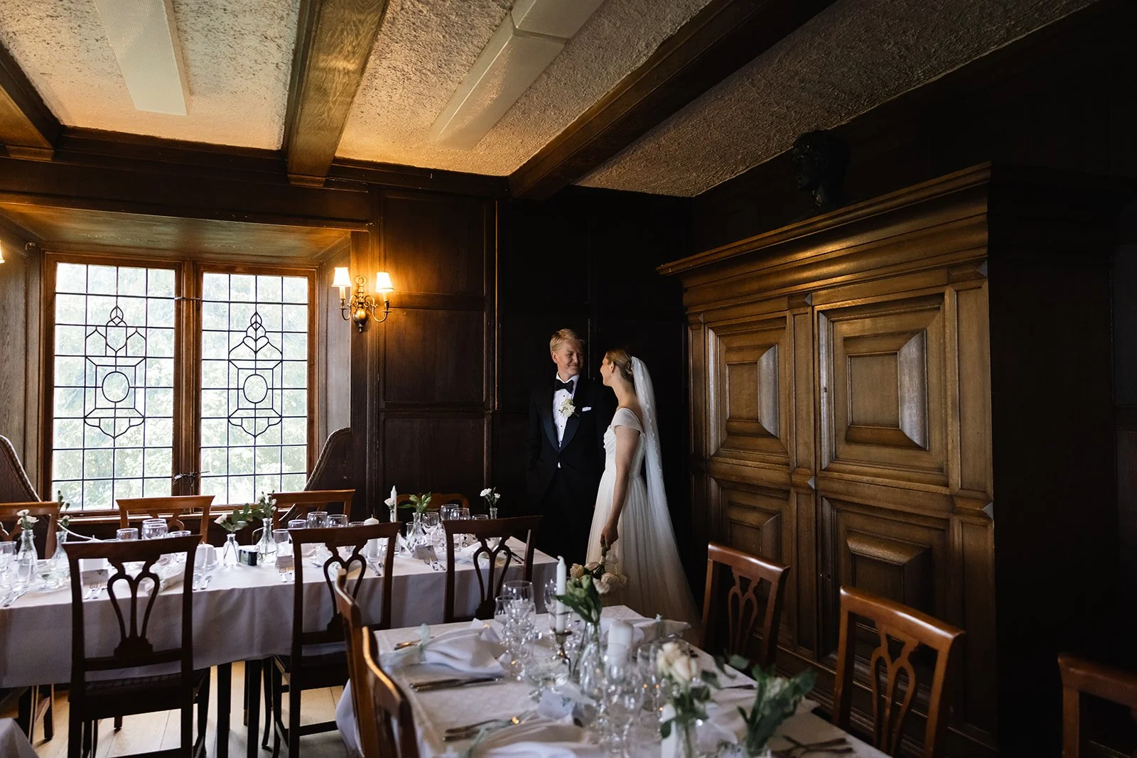 En brud og en brudgom står og ser på hverandre i et rom med tre vinduer, mørk panelvegg og en stor trebord dekket med hvit duk, glass og blomster. Bruden har hvit kjole og brudekveil, mens brudgommen har svart dress.
