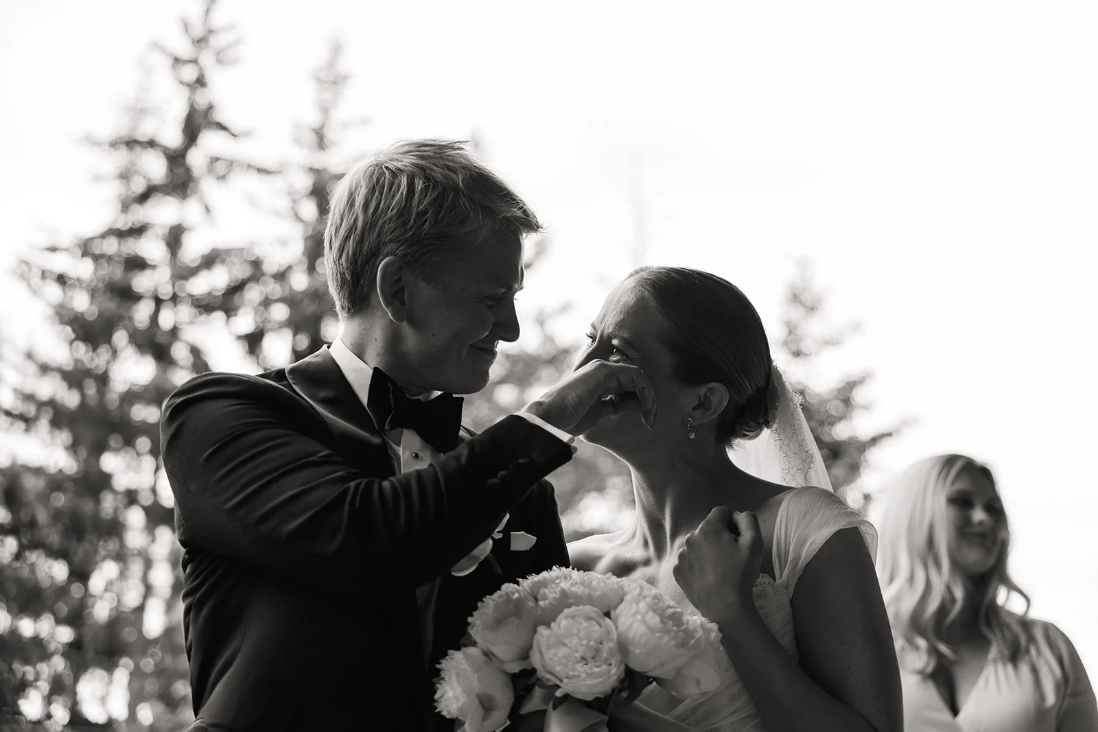 Et svart-hvitt bilde av et bryllup der en mann i dress og sløyfe og en kvinne i brudekjole er emosjonelle, med mannen som tørker tårene til kvinnen. En kvinne i bakgrunnen smiler.