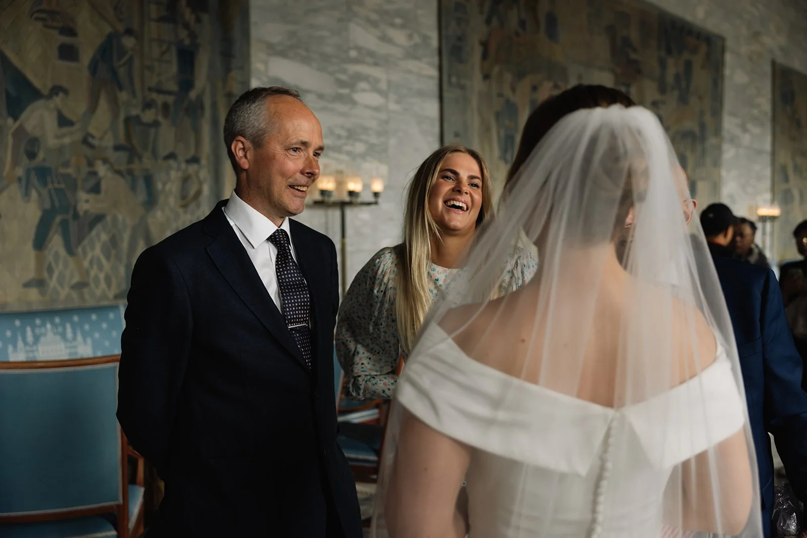 En brud med hvit off-shoulder kjole og slør som snakker med tre personer, en eldre mann i dress og to unge kvinner, i et kunstnerisk interiør med veggmalerier og levende lys.