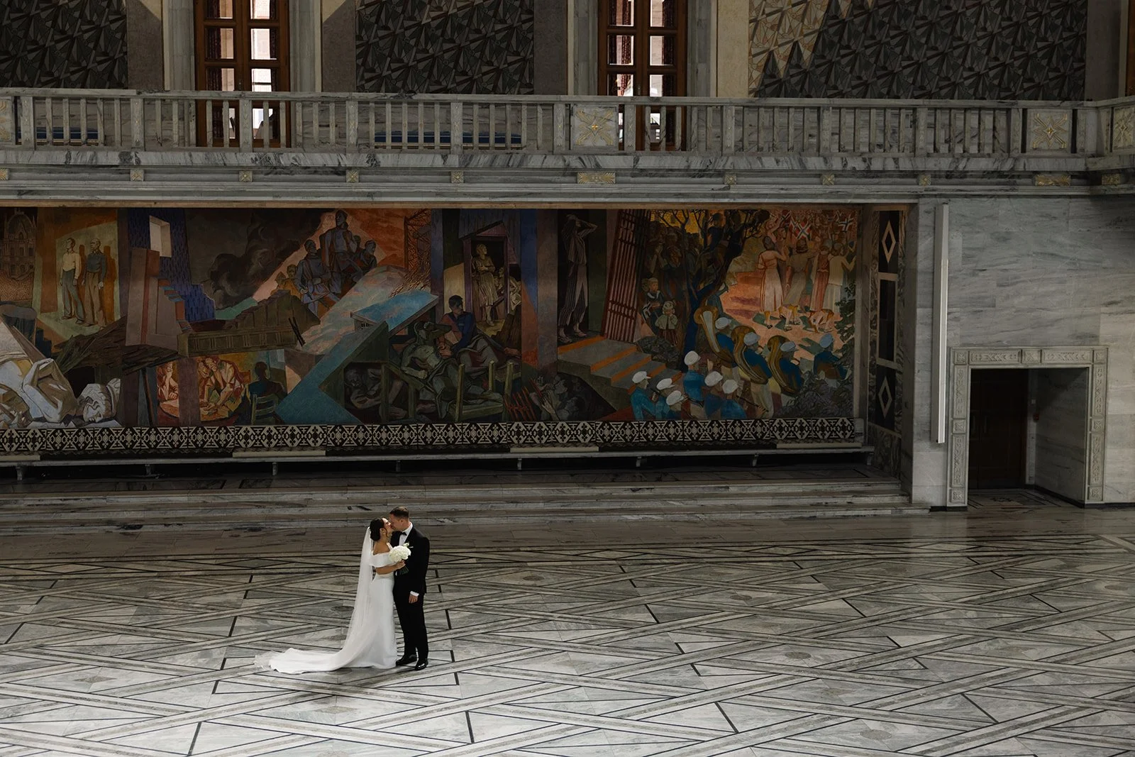 Et brudepar står og kysser i et stort, tradisjonelt bygg med marmorgulv og et stort maleri på veggen bak dem.
