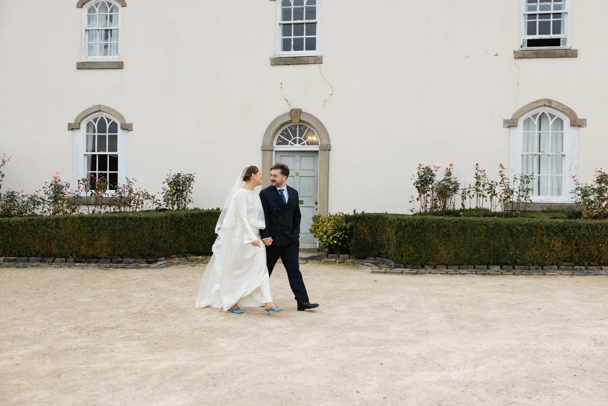 Et brudepar går hånd i hånd foran en hvit bygning med flere vinduer og en dør, omgitt av busker og blomster.