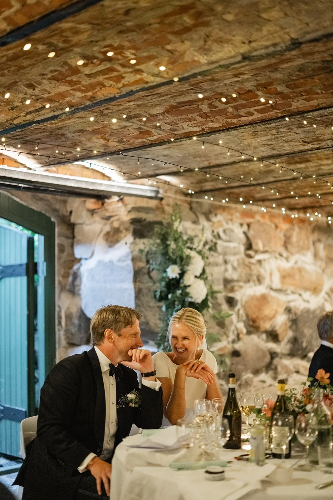 Et lite selskap som feirer, med to personer i fokus som smiler og snakker. De er kledd pent, med en bordplate som har vin, glass, blomster og lys, og det er en rustikk steinvegg og treverk i bakgrunnen, pyntet med festlys.