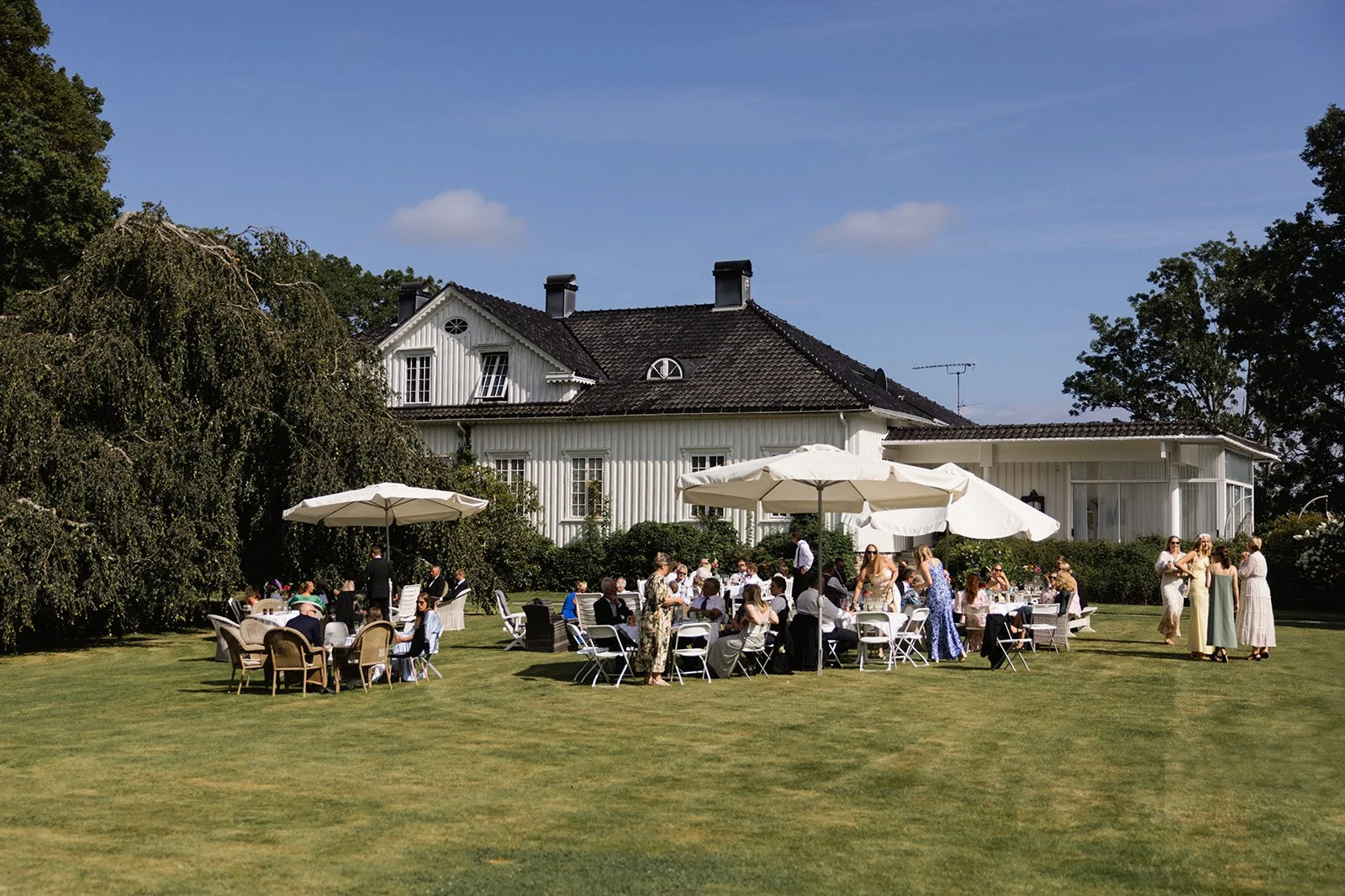 Utendørs bryllup med pyntede bord, parasoller og gjester i hage foran en hvit bolig.