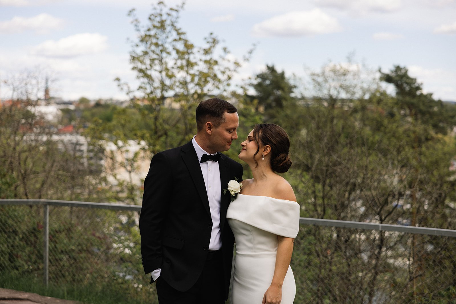 Et bryllupspanel hvor en mann i tuxedo og en kvinne i en off-shoulder hvit kjole står nært sammen, med en bakgrunn av trær og en uklar bylandskap.