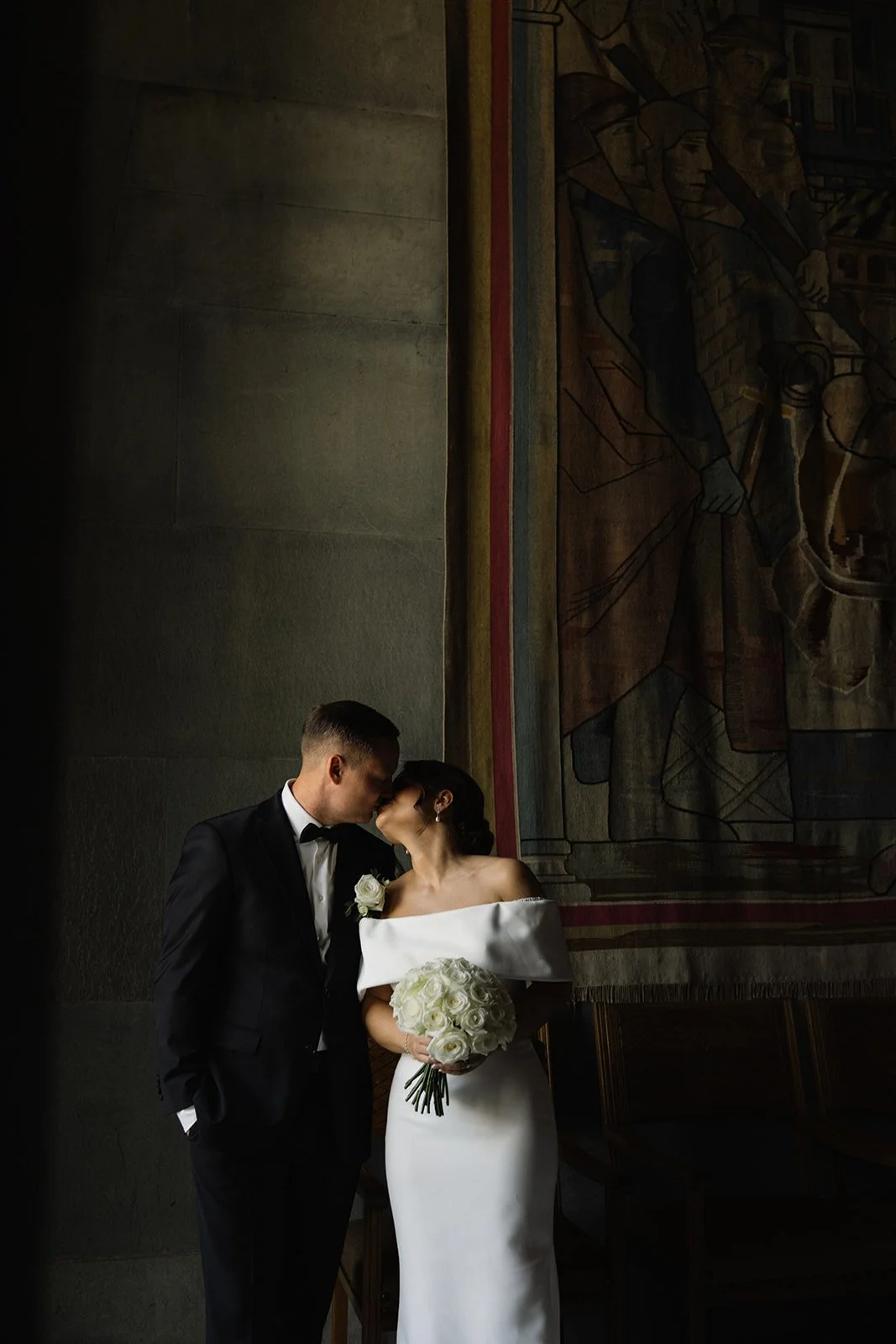Et bryllupsbilde av en kledd brud og brudgom som kysser i et mørkt rom med en stor veggkunst som bakgrunn.