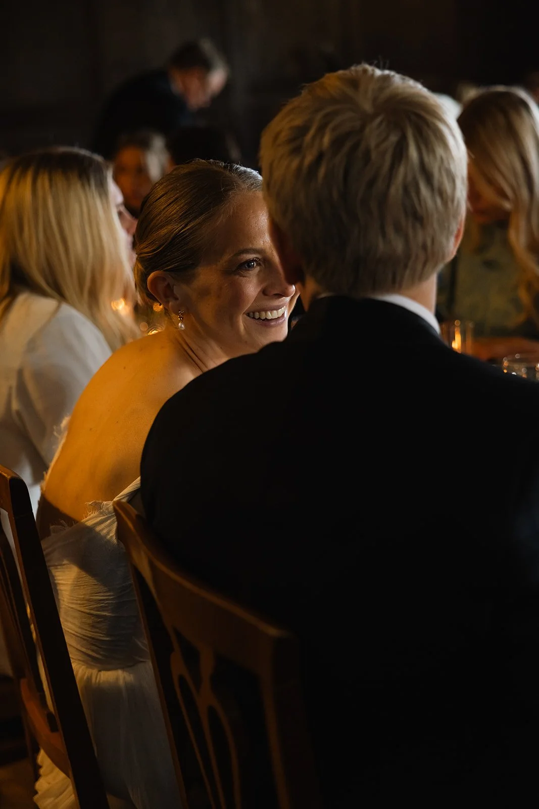 Et gruppe mennesker sitter i en mørk restaurant, og en kvinne smiler mens hun snakker med en mann med blond hår. Venninner eller kolleger er synlige rundt dem.