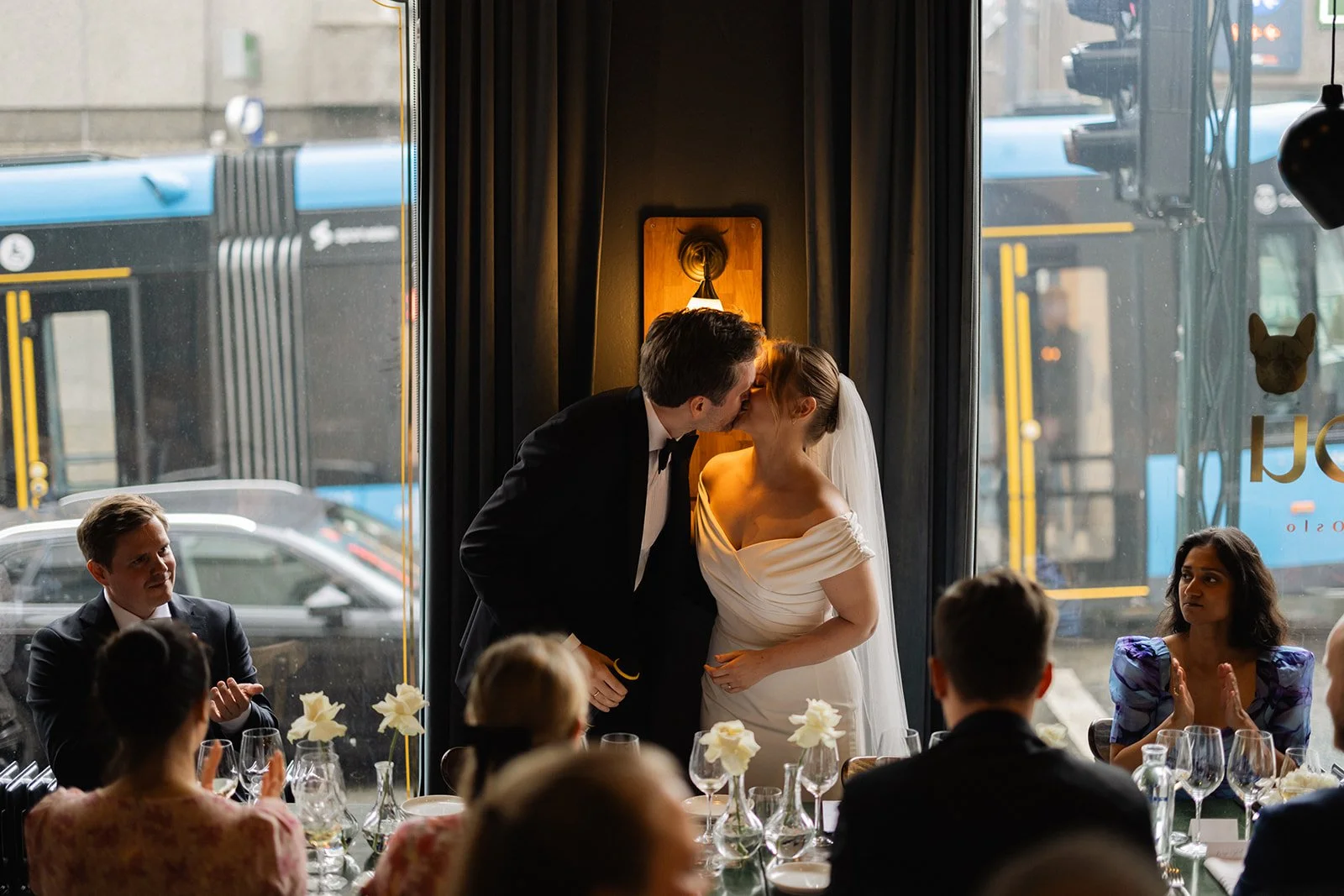Et brudepar kysser foran et bord med brude- og brudgom samt gjester under deres bryllupsseremoni i en innendørs restaurant.