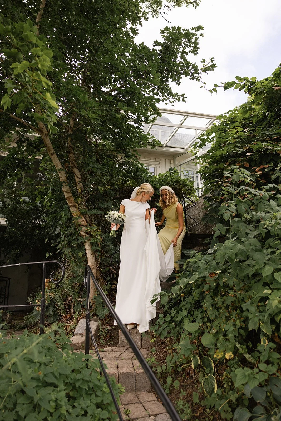 To kvinner i brudekjoler går nedover trapp i en hage med frodig grønt. Den ene kvinnen holder en blomsterbukett.