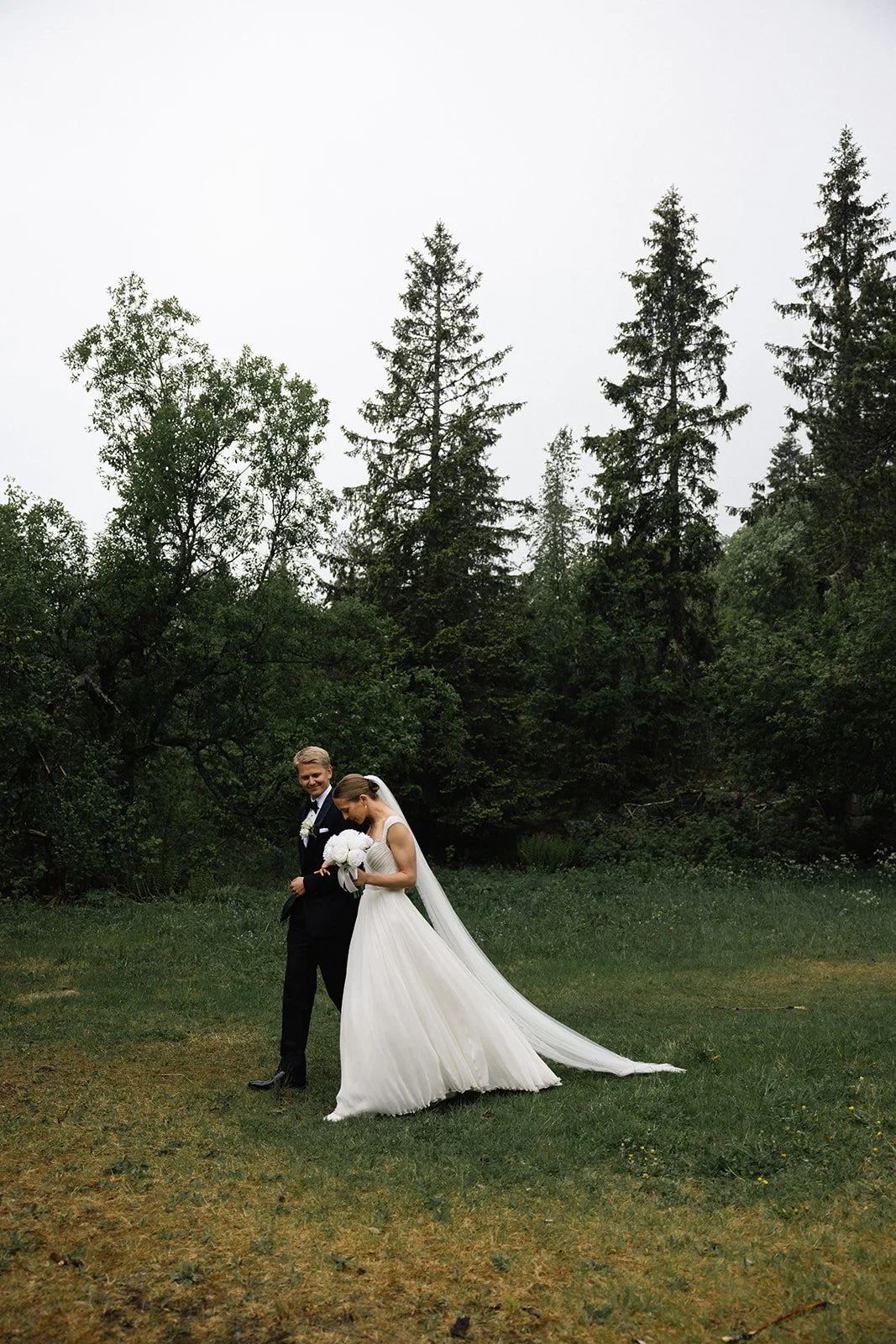Et brudepar går hånd i hånd i en grønn skog, med kvinnen i brudekjole og mannen i svart dress, begge glade.