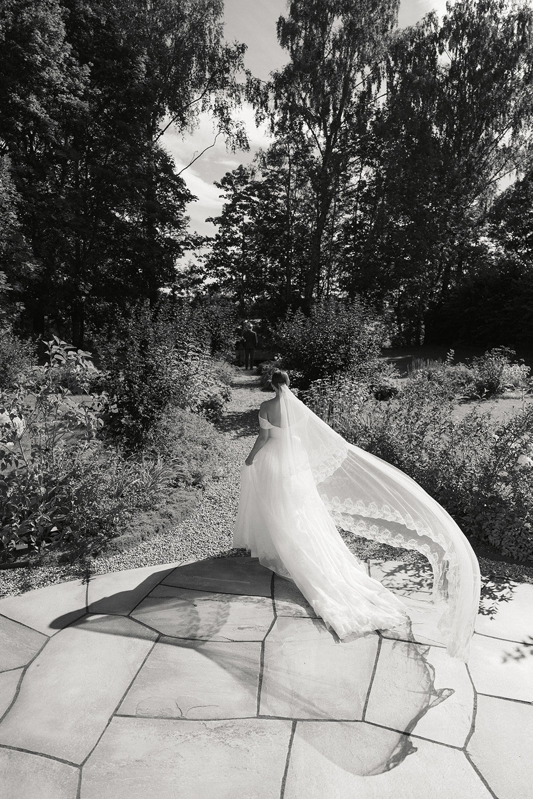 En kvinne i en bryllupskjole går på en sti i en hage med trær og busker, i svart-hvitt.